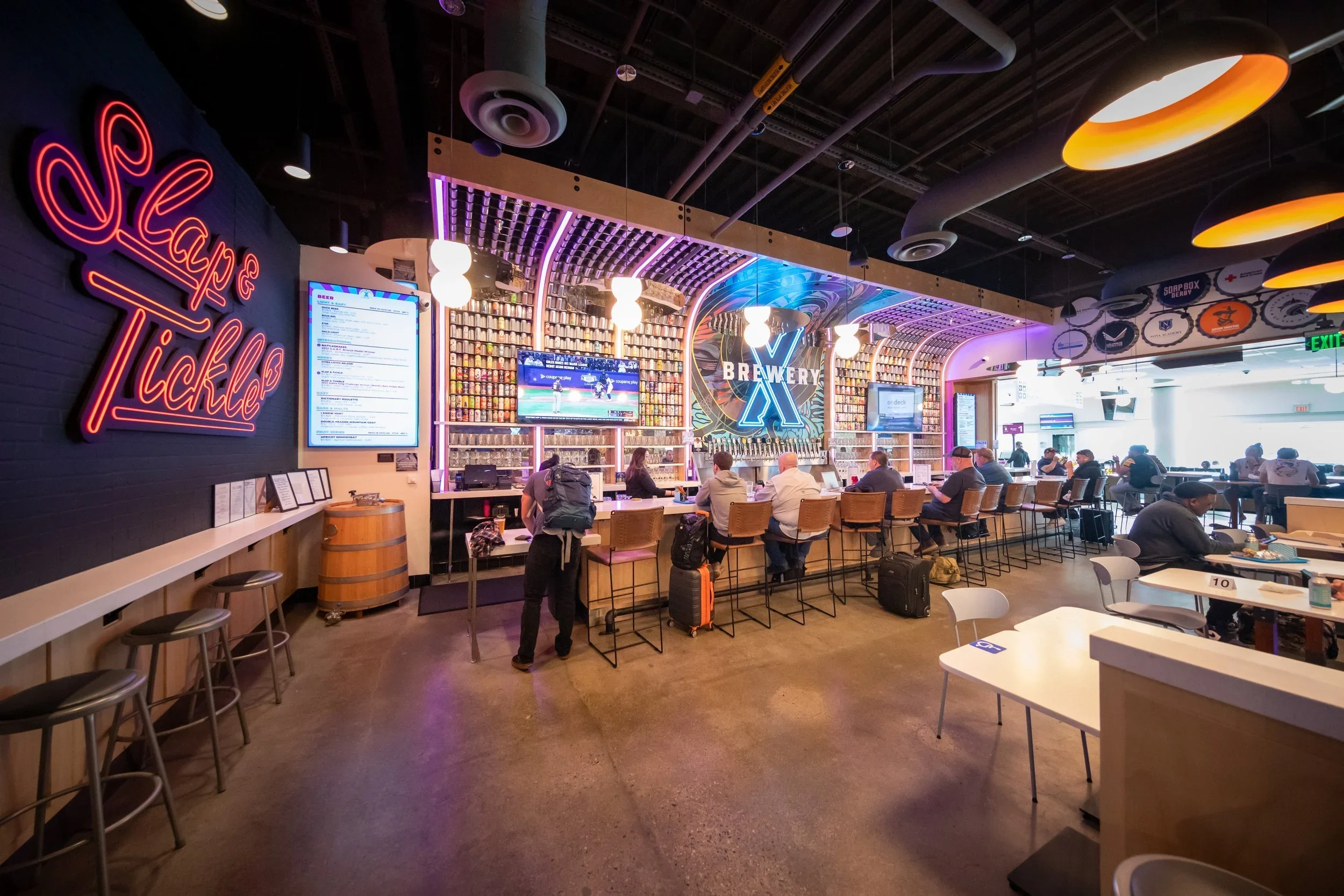 The Slaps & Tickles bar inside Ontario International Airport featuring a Brewery X tap wall, neon signage, and travelers seated at the counter with luggage, set against a vibrant lit backdrop of stacked beer cans.