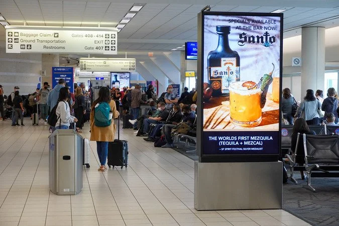 A digital display advertising Santo Tequila, the world's first Mezquila, positioned in a busy ONT terminal corridor near gate seating areas, with travelers passing through in the background.