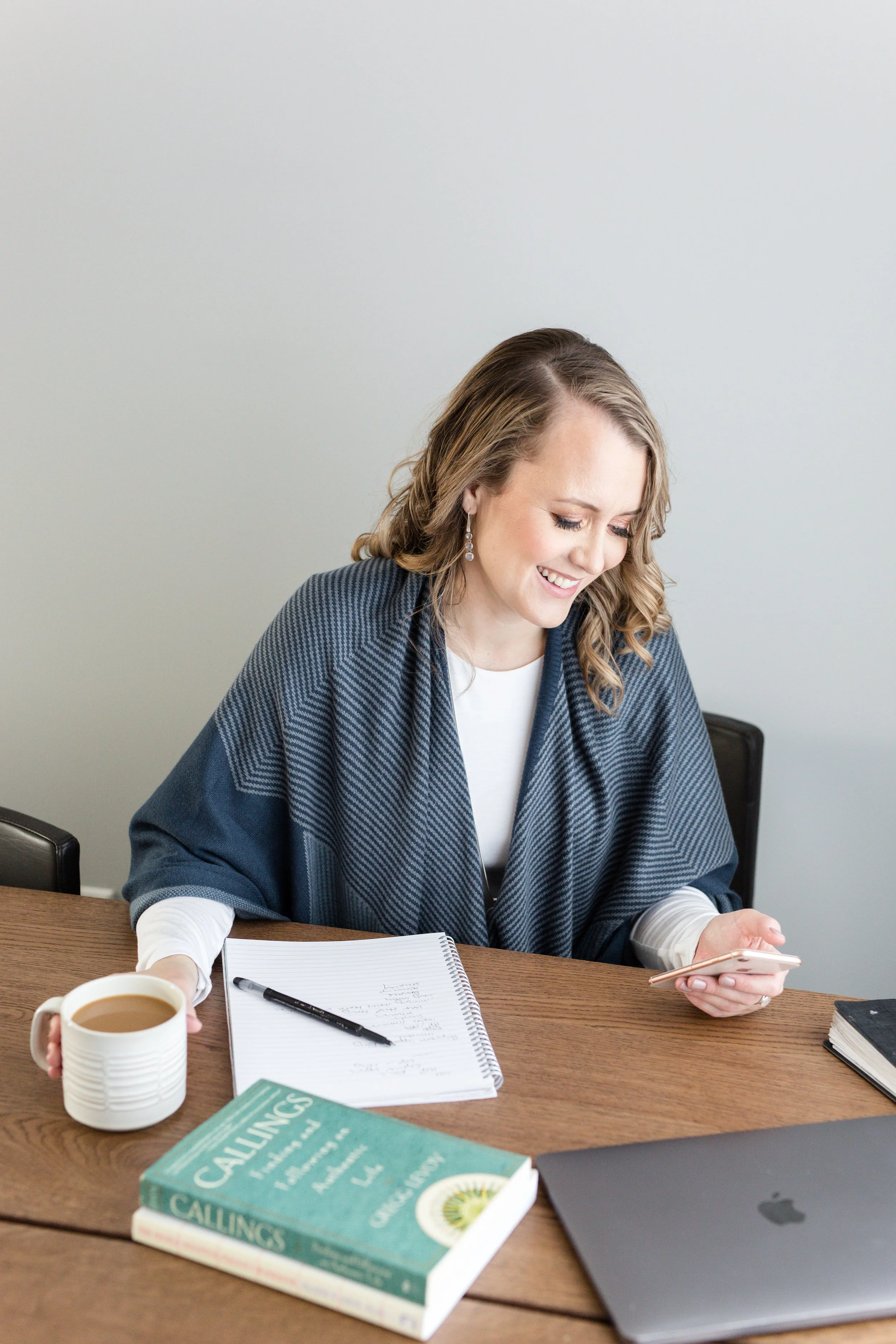 A woman with curly blonde hair wearing earrings and a blue blazer is sitting at a wooden table, smiling and looking at her phone. There is an open notebook, a pen, a book titled 'Callings', a laptop, a black notebook, a cup of coffee, and other papers on the table.