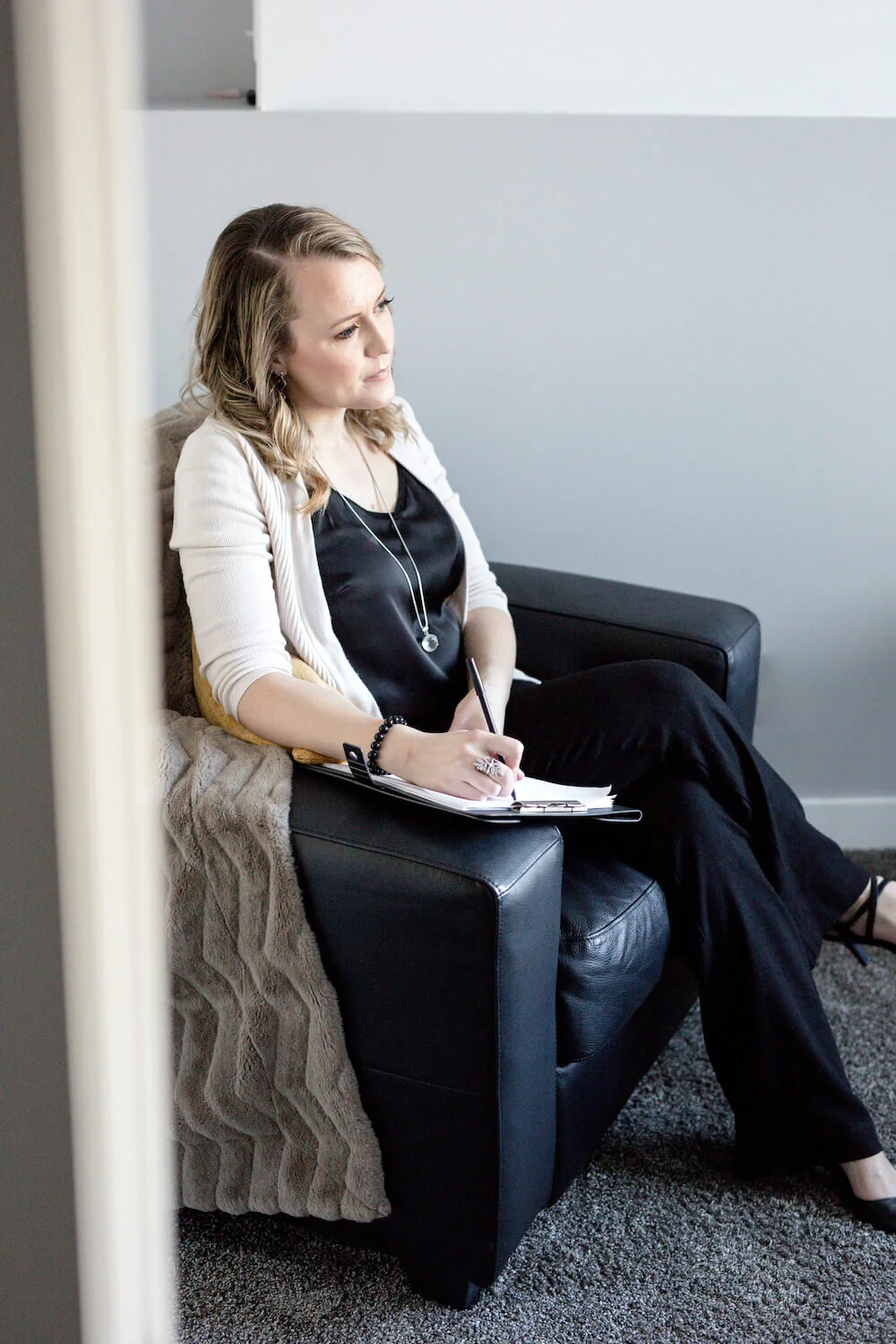 A woman sitting on a black leather chair in a modern room, taking notes on a clipboard, wearing a black dress, cream-colored cardigan, and black shoes, with a blanket draped over the armrest.
