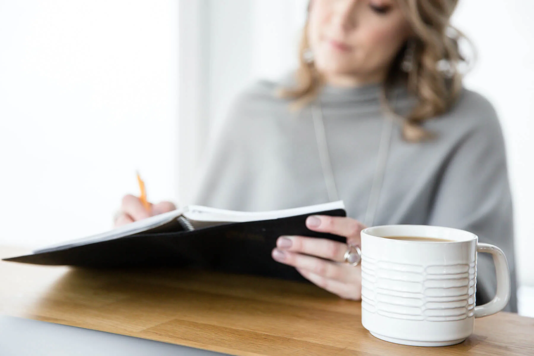Woman in gray hoodie sitting at a wooden desk, writing in a notebook, with a cup of coffee nearby.