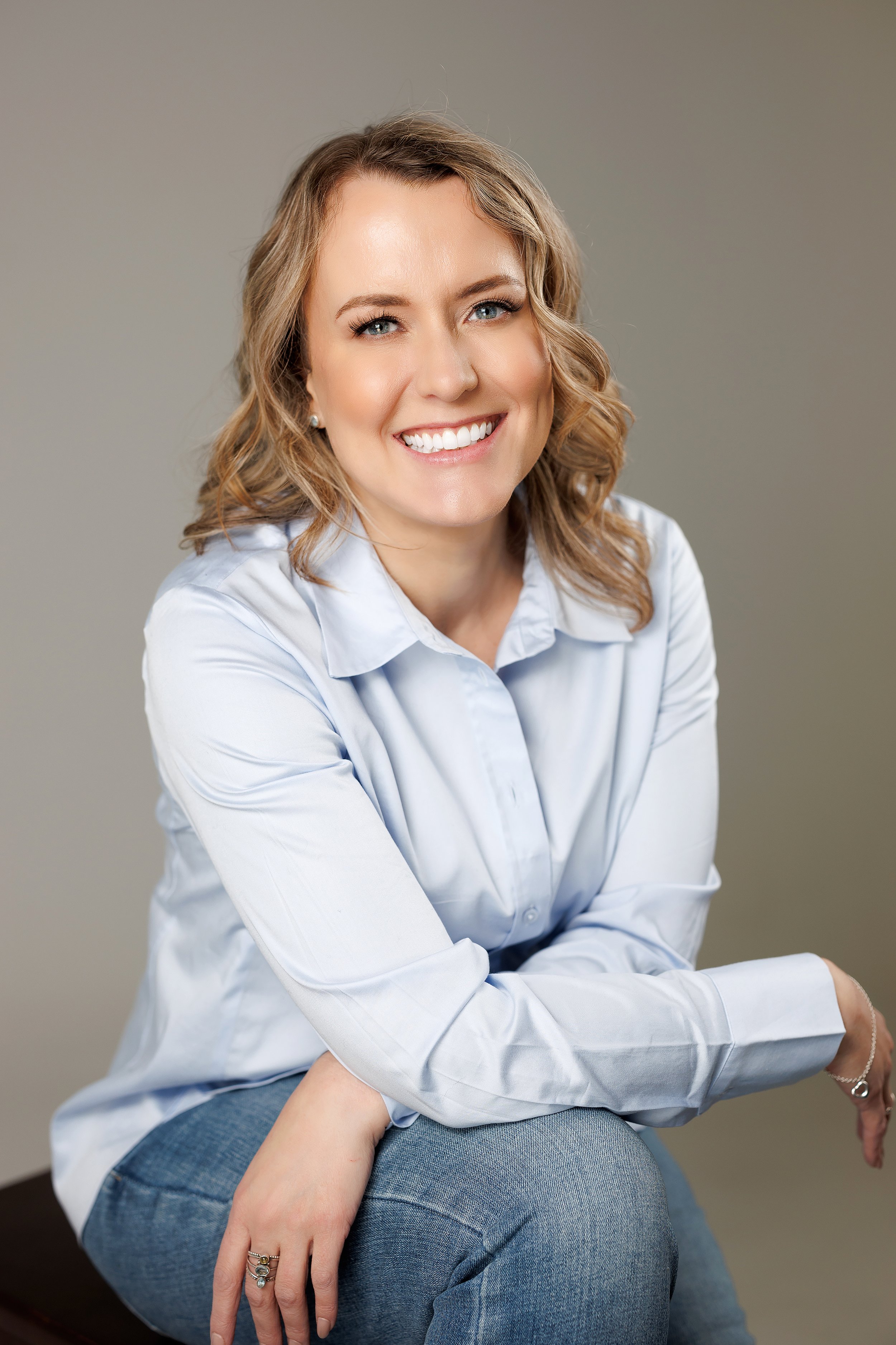 A woman with blonde hair, blue eyes, and a bright smile, wearing a white button-up shirt and blue jeans, sitting with her arms crossed on her lap, against a neutral background.
