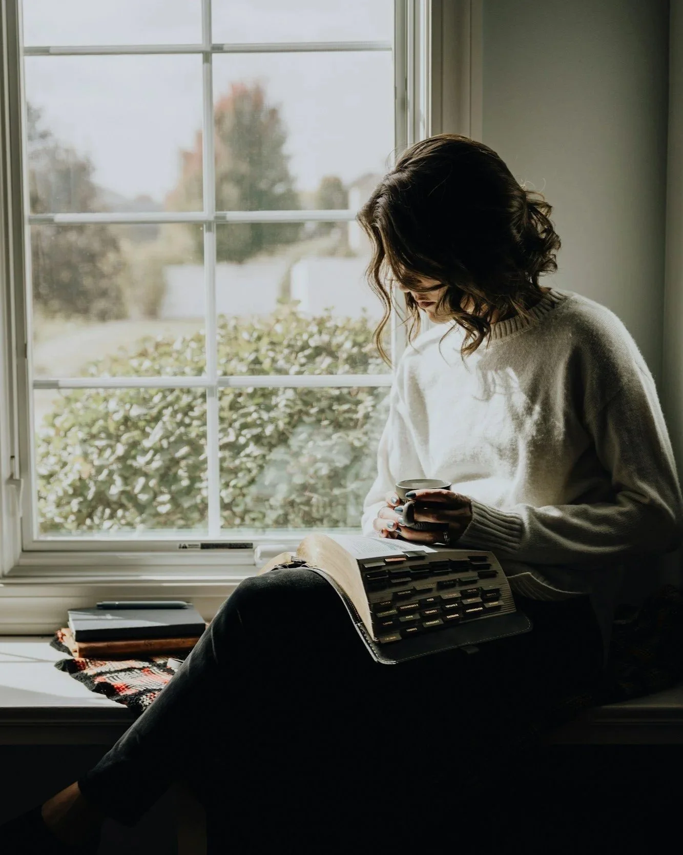 A woman with dark, curly hair sitting on a windowsill, holding a mug, with a book and a calculator on her lap. The window shows bushes and trees outside.