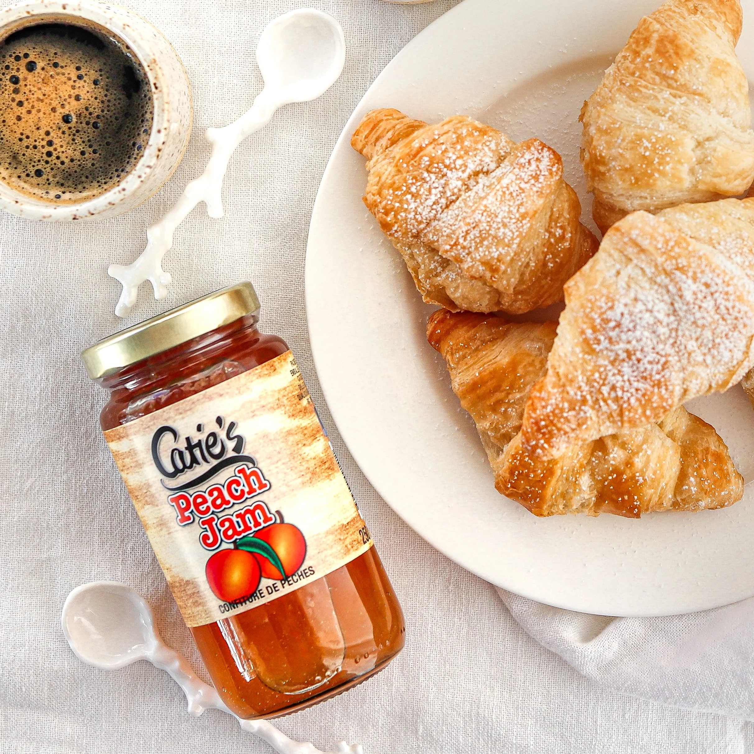 A plate of croissants, a jar of peach jam, a cup of black coffee, and a white decorative spoon on a light tablecloth.