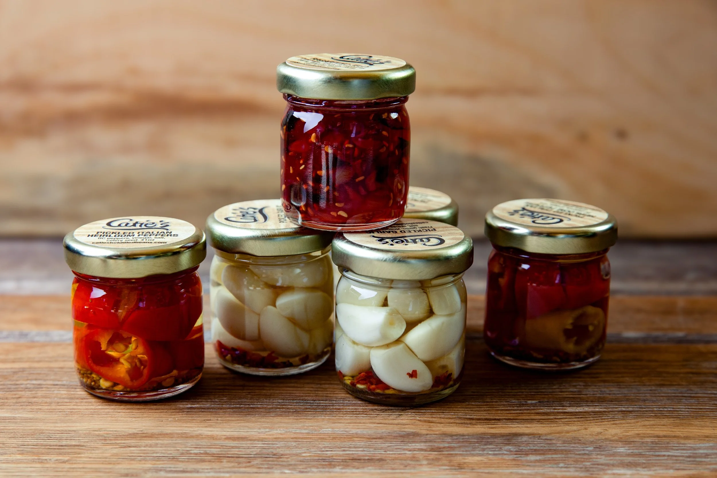 Six small glass jars with gold lids, containing various pickled vegetables, arranged on a wooden surface with a wooden background.