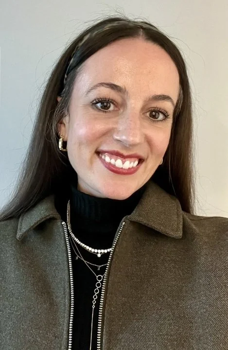 A woman with long brown hair, smiling, wearing a black turtleneck, layered necklaces, gold hoop earrings, and a brown zip-up jacket.