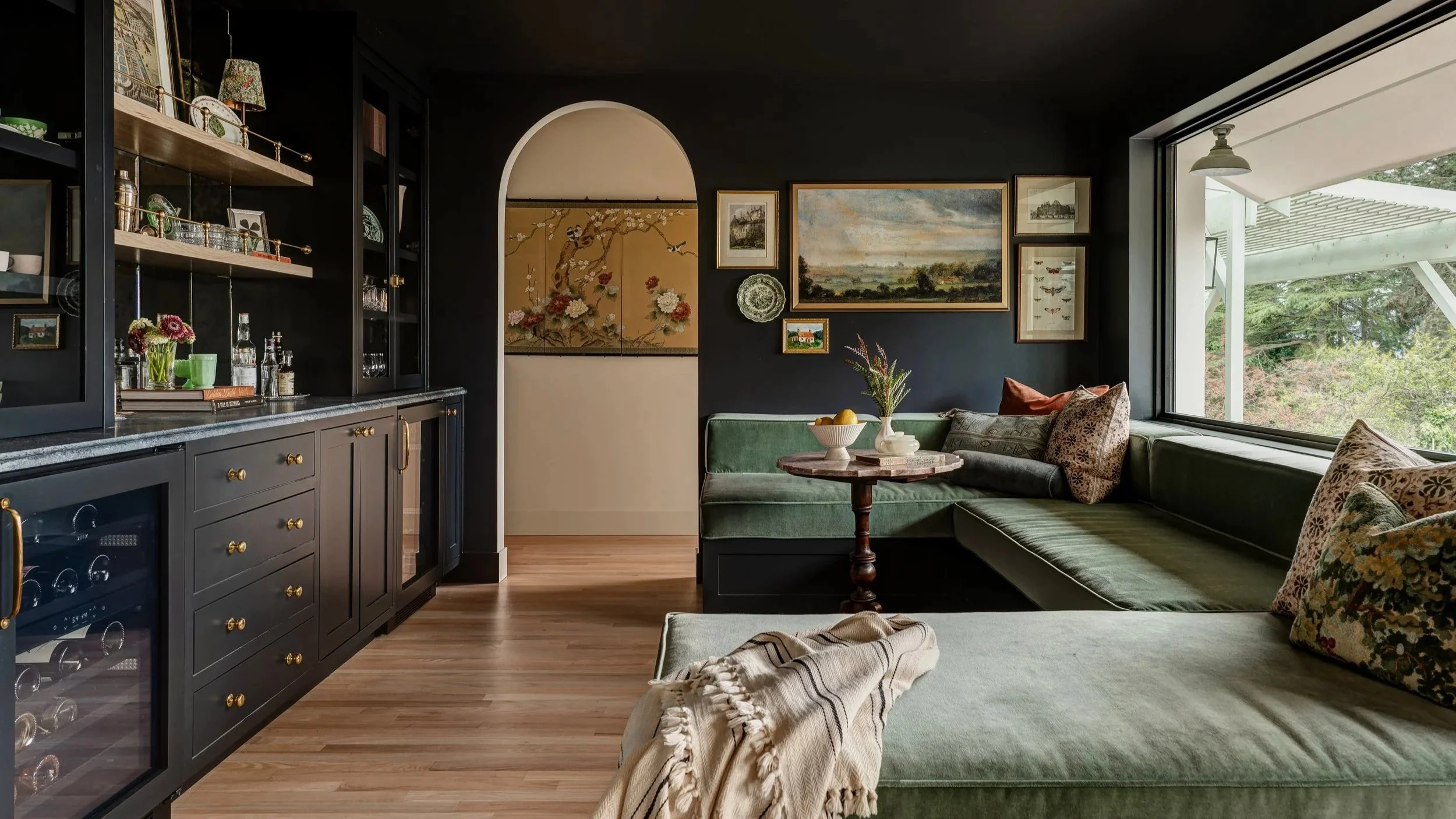 Living room with black walls, a large window, green sectional sofa with patterned pillows, and a round wooden table with a bowl of fruit and a vase of flowers. Artwork and decorative plates hang on the wall, and a black cabinet with glass doors displ