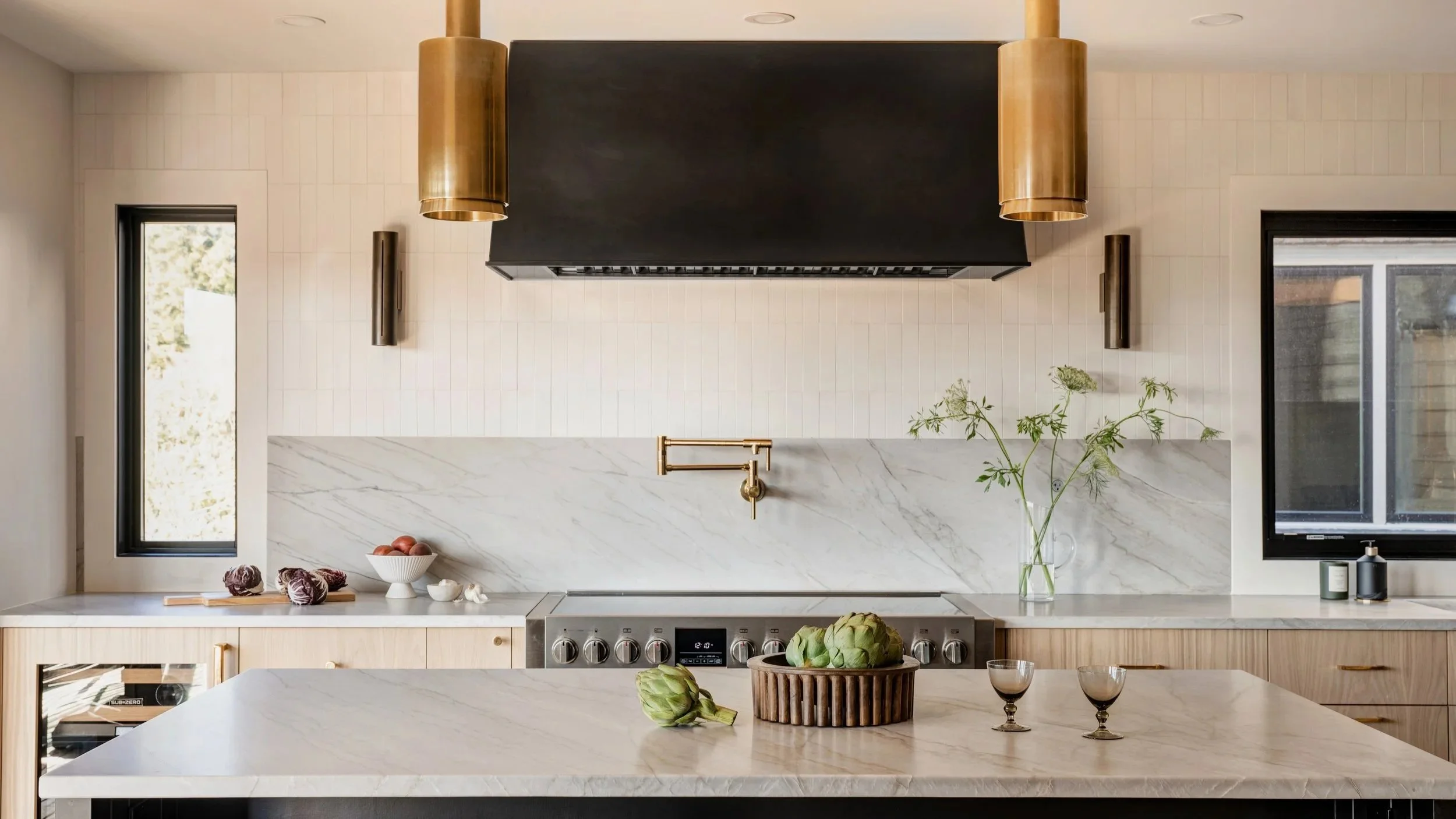 Modern kitchen with white marble island and backsplash, black vent hood, wooden cabinetry, gold accents, and large windows with greenery outside.