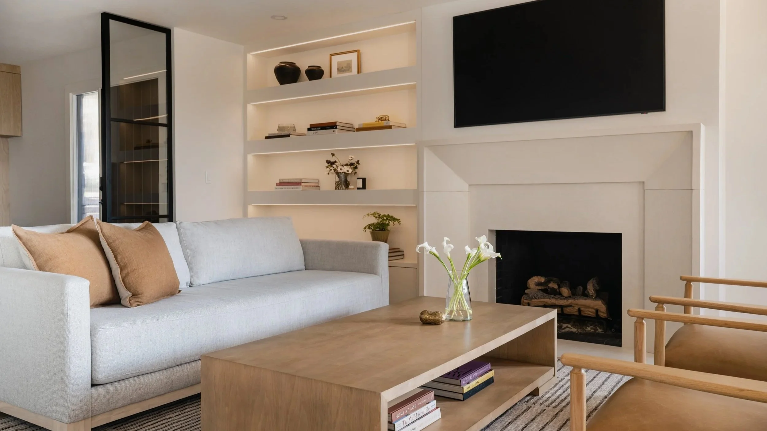 Modern living room with a light gray sofa, wooden coffee table, and a fireplace with a large black TV above it. Shelves with decor and books are on the wall. There are two tan chairs and a vase with white flowers on the coffee table.