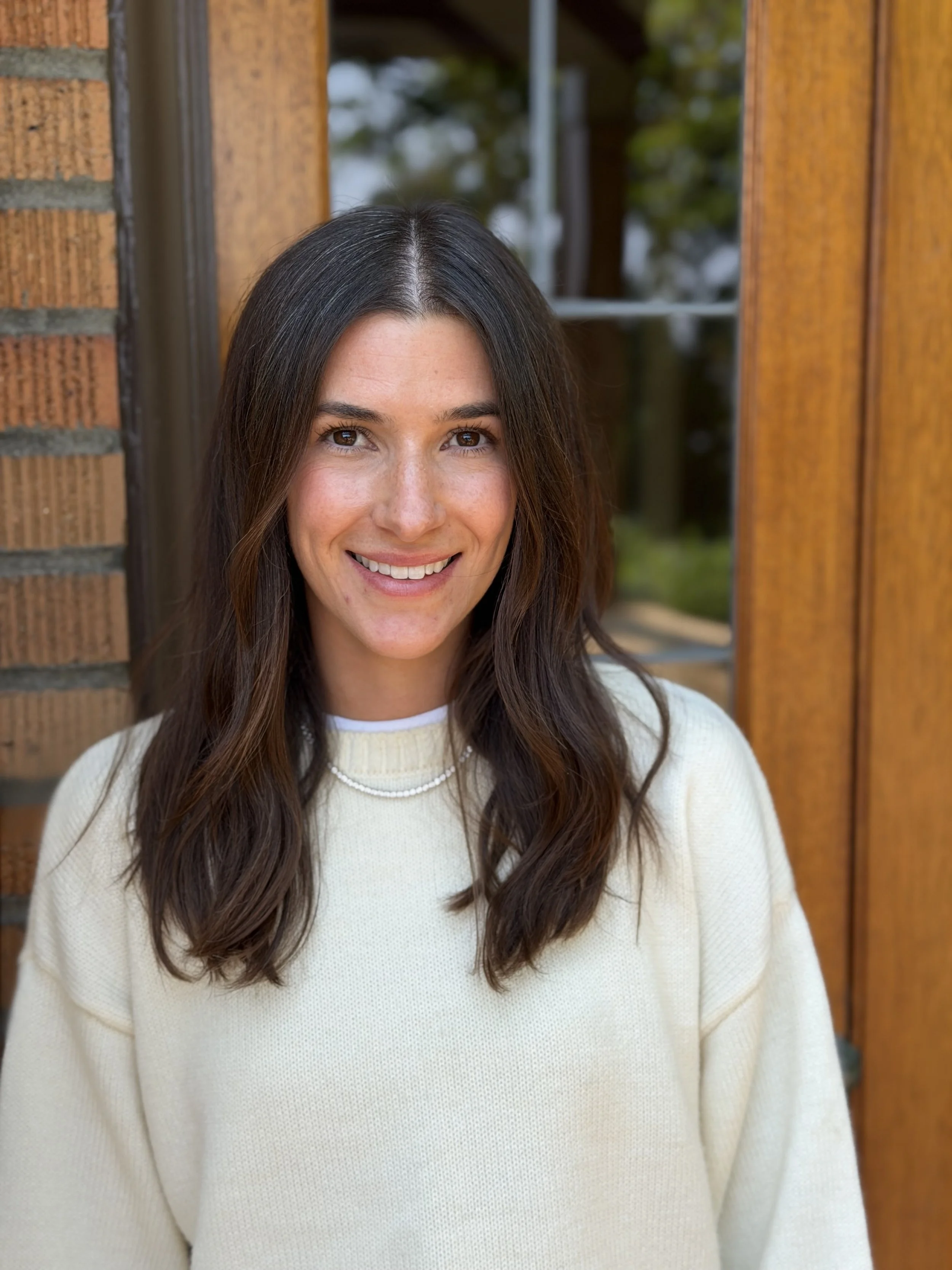A woman with long dark hair smiling, standing outside near a brick wall and wooden window frame.