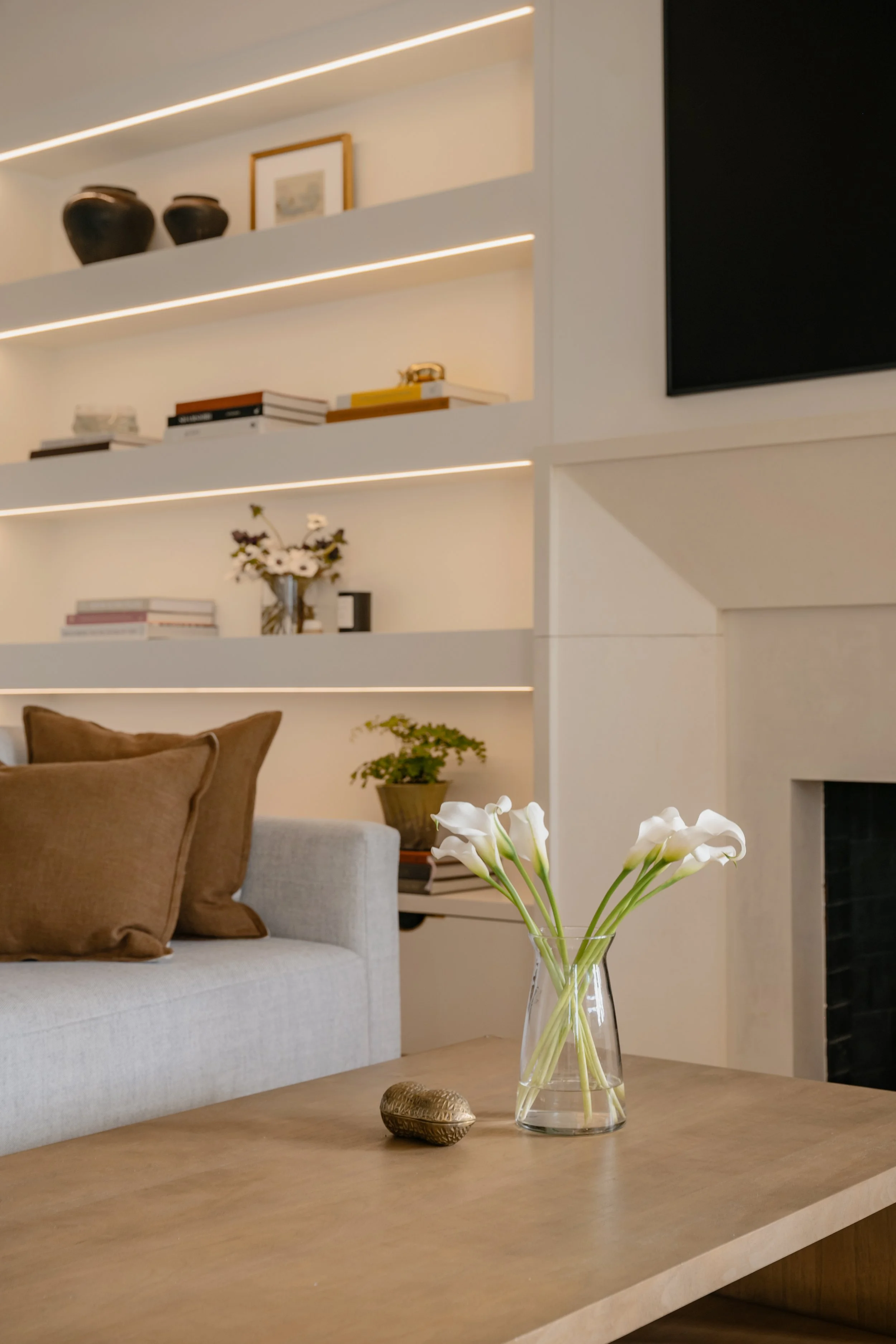Modern living room with a beige sofa, decorative pillows, a wooden coffee table with a glass vase of white calla lilies, a fireplace, and built-in shelves with books, vases, and decorative objects.