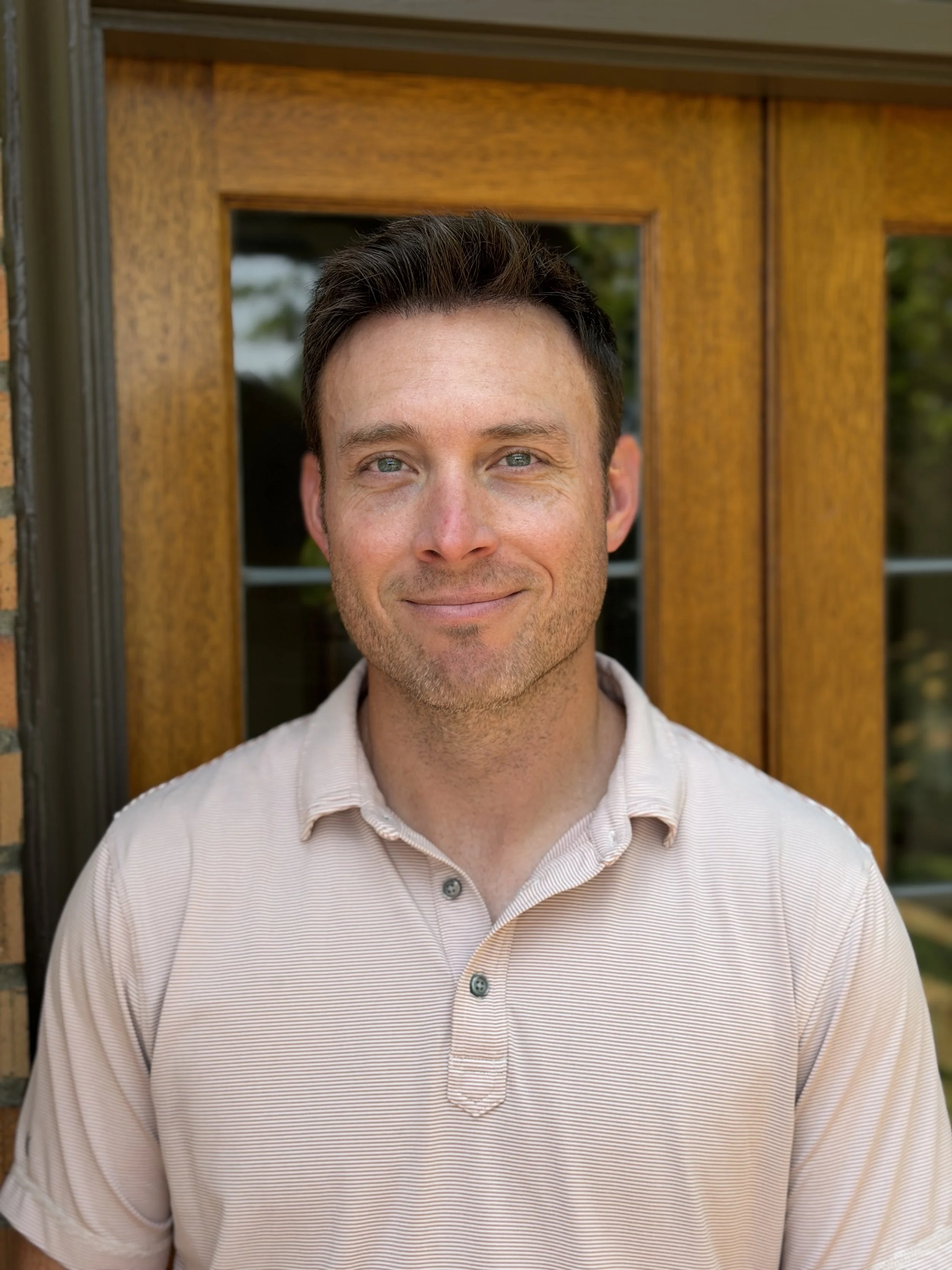 A close-up portrait of a smiling man with short dark hair, blue eyes, and light facial hair, wearing a gray shirt against a plain background.