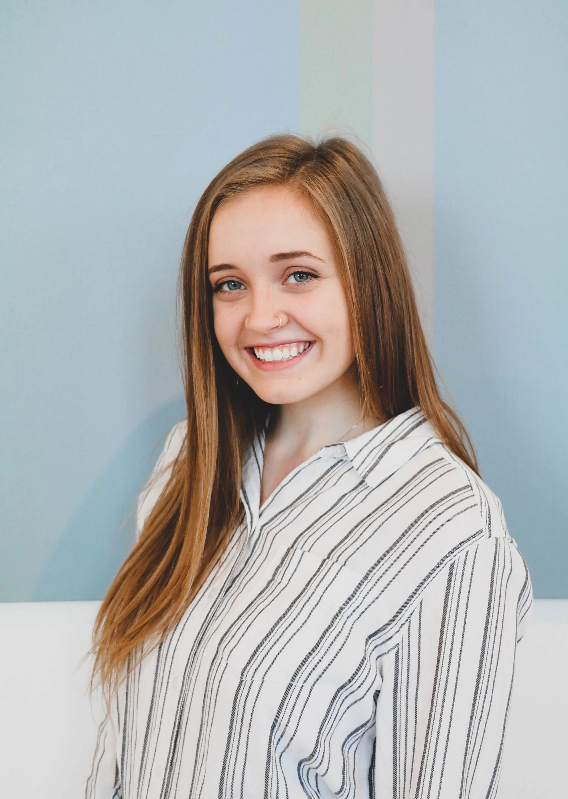 Smiling young brunette woman in striped button-down shirt with blue background.