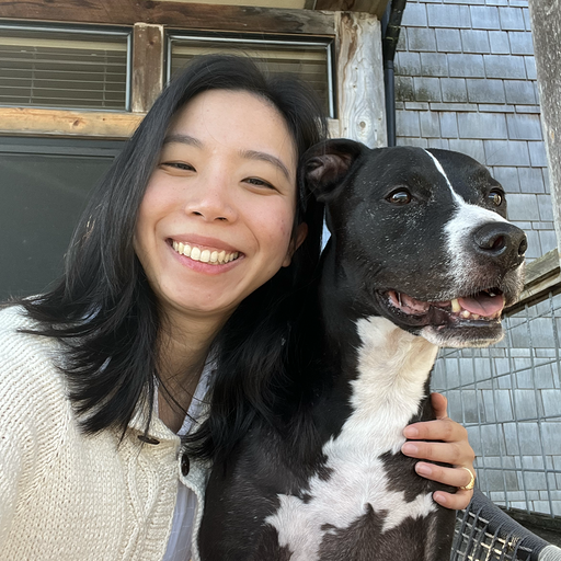 A young woman smiling and taking a selfie with her black and white dog outside near a house with wooden and shingled walls.
