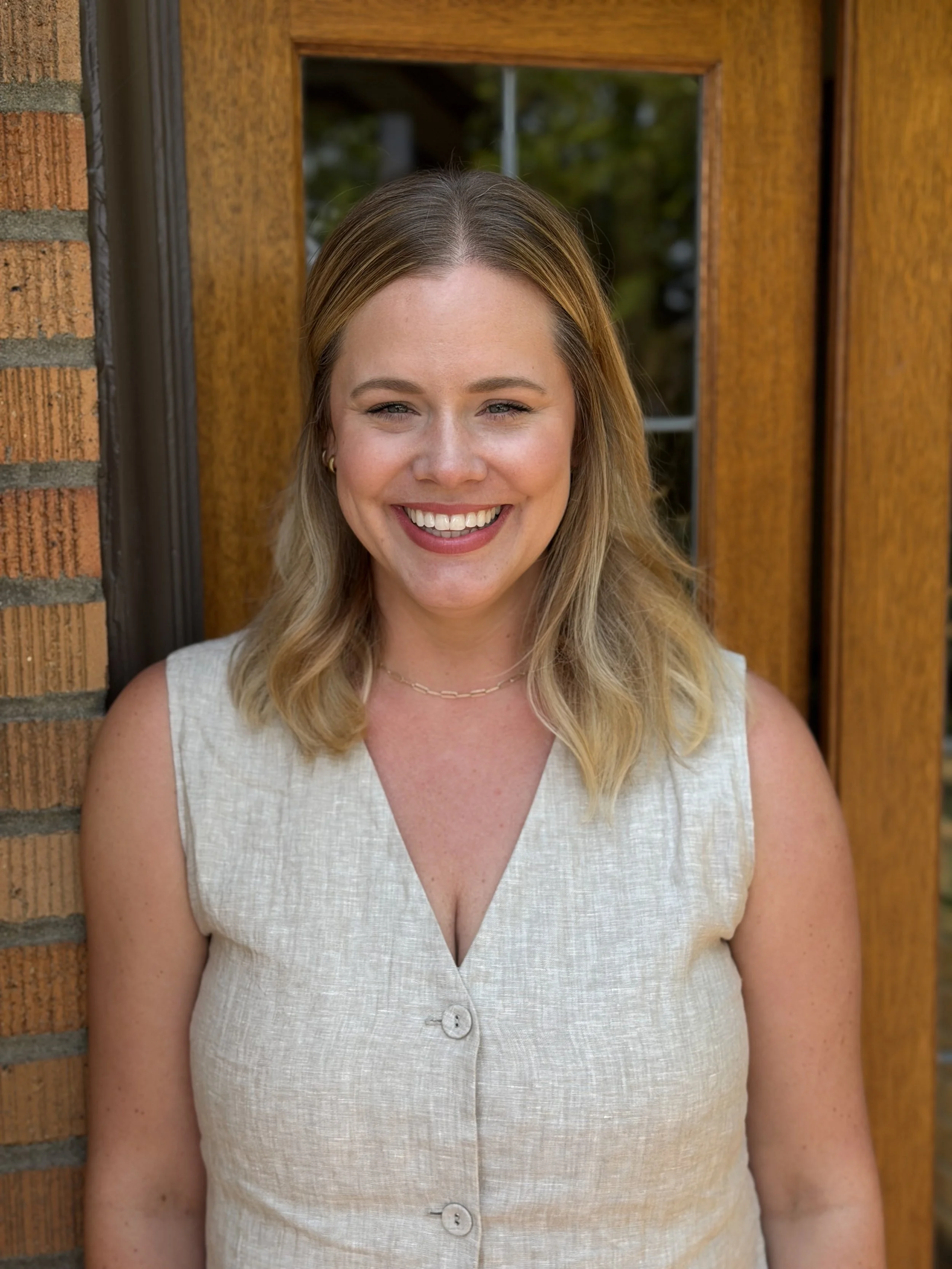 A woman with shoulder-length blonde hair smiling outdoors with green foliage in the background.