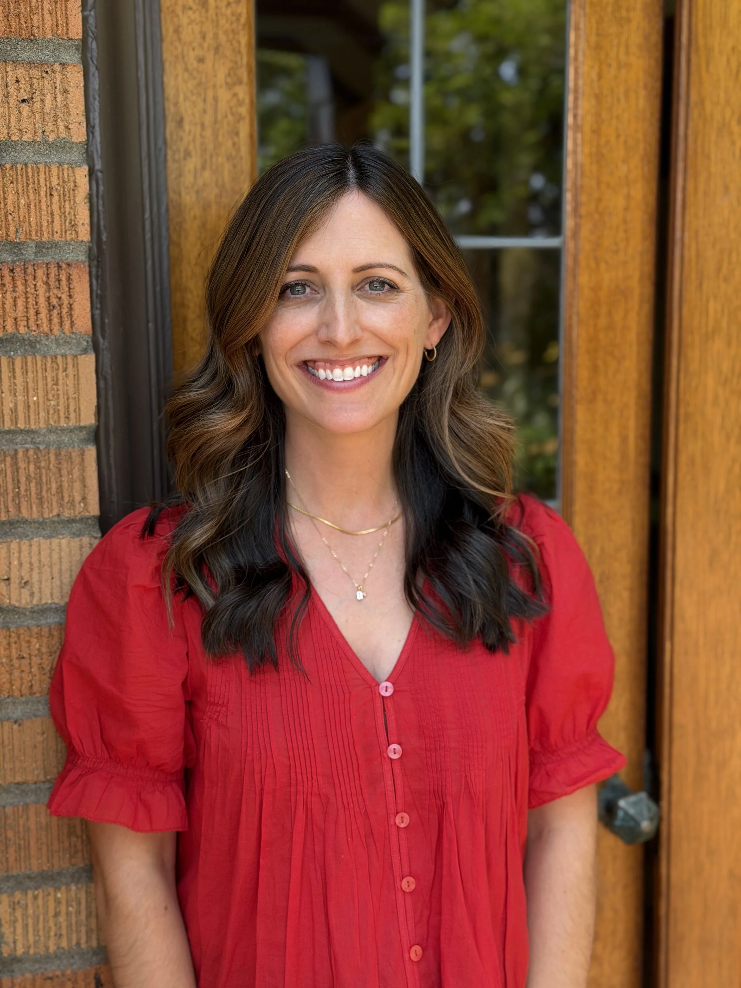 A woman with shoulder-length, wavy brown hair, smiling outdoors with colorful flowers in the background.