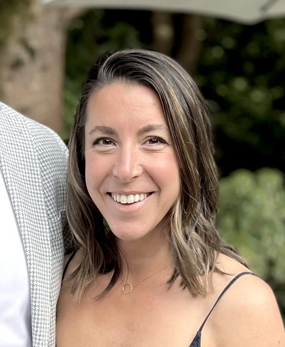 A woman with shoulder-length brown hair, smiling outdoors, wearing a black dress with thin straps and a gold necklace, with a blurred green background.