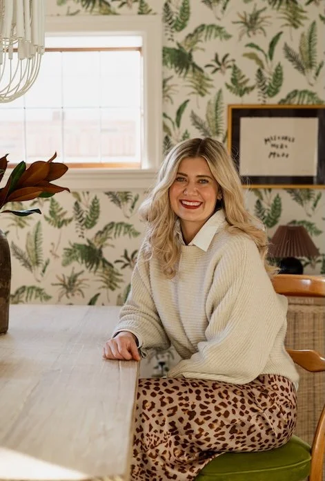 Blonde woman with red lipstick sitting in dining room with botanical wallpaper