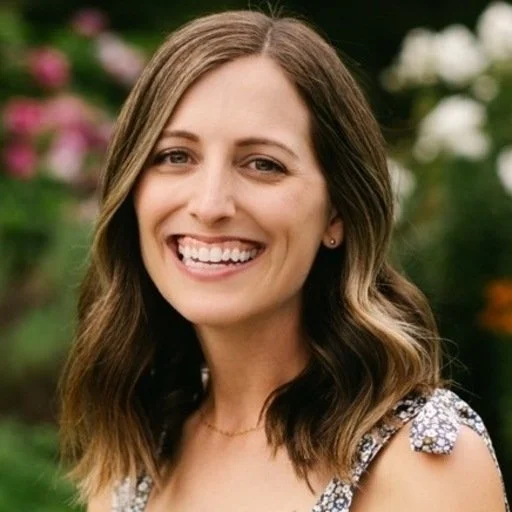 A woman with shoulder-length, wavy brown hair, smiling outdoors with colorful flowers in the background.