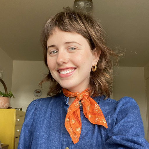 A young woman smiling with short brown hair, wearing a blue top and an orange bandana around her neck, in a room with a ceiling light and some furniture in the background.