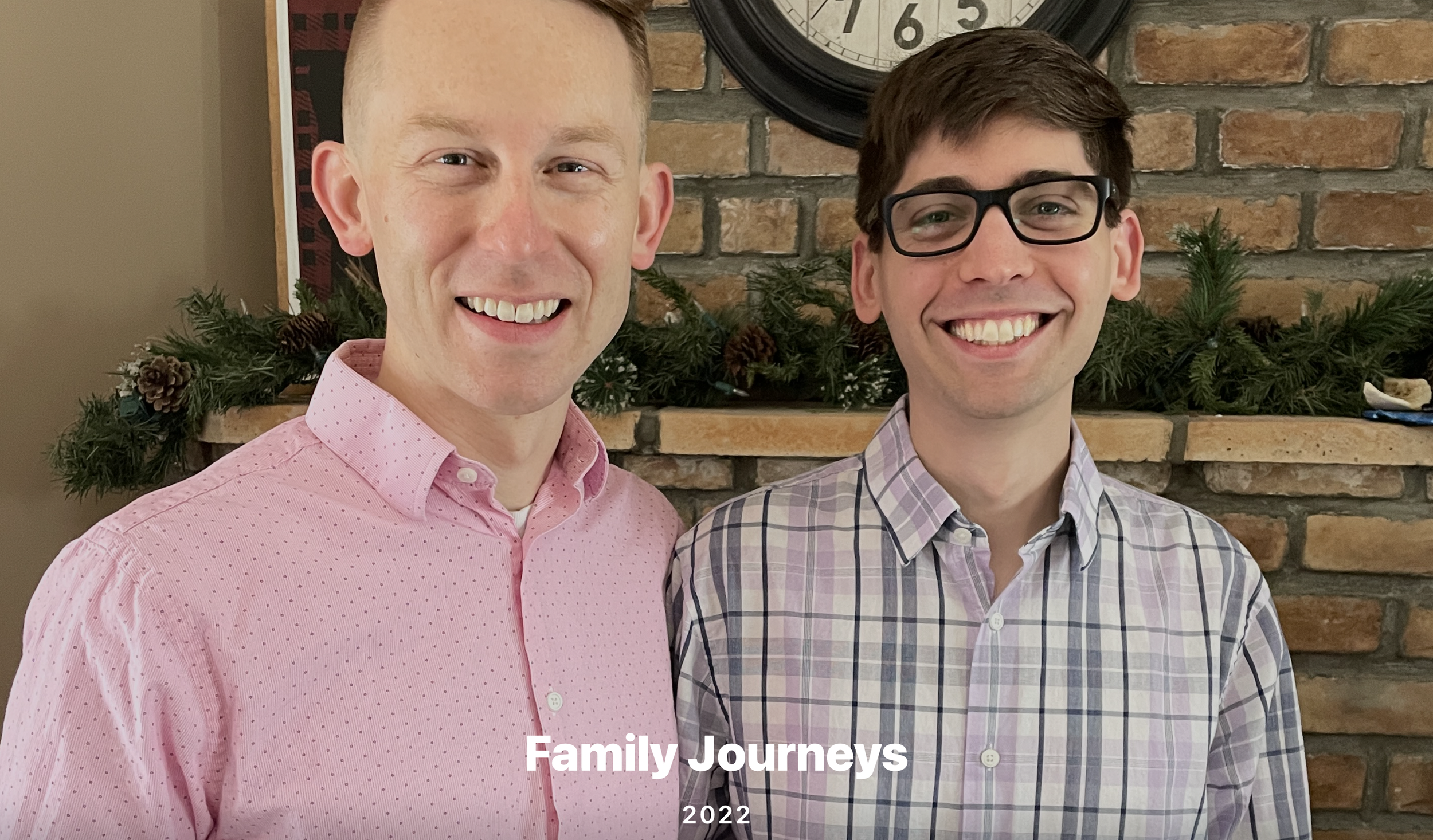 Two smiling men standing in front of a brick fireplace decorated with greenery and pinecones, with a clock hanging above them. The man on the left is wearing a pink dress shirt, and the man on the right is wearing glasses and a plaid button-up shirt. Text at the bottom reads 'Family Journeys 2022'.