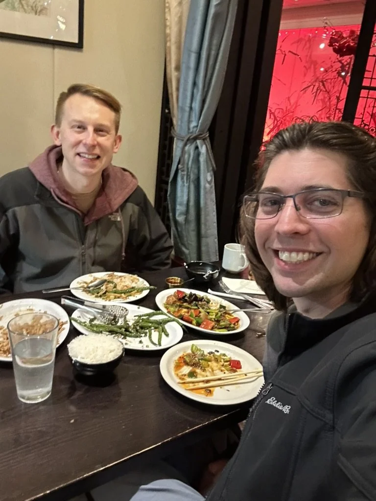 Two young men are sitting at a dinner table with Asian cuisine dishes, including rice, green beans, vegetables, and seafood, in a restaurant with a decorative curtain and red lighting outside a window.