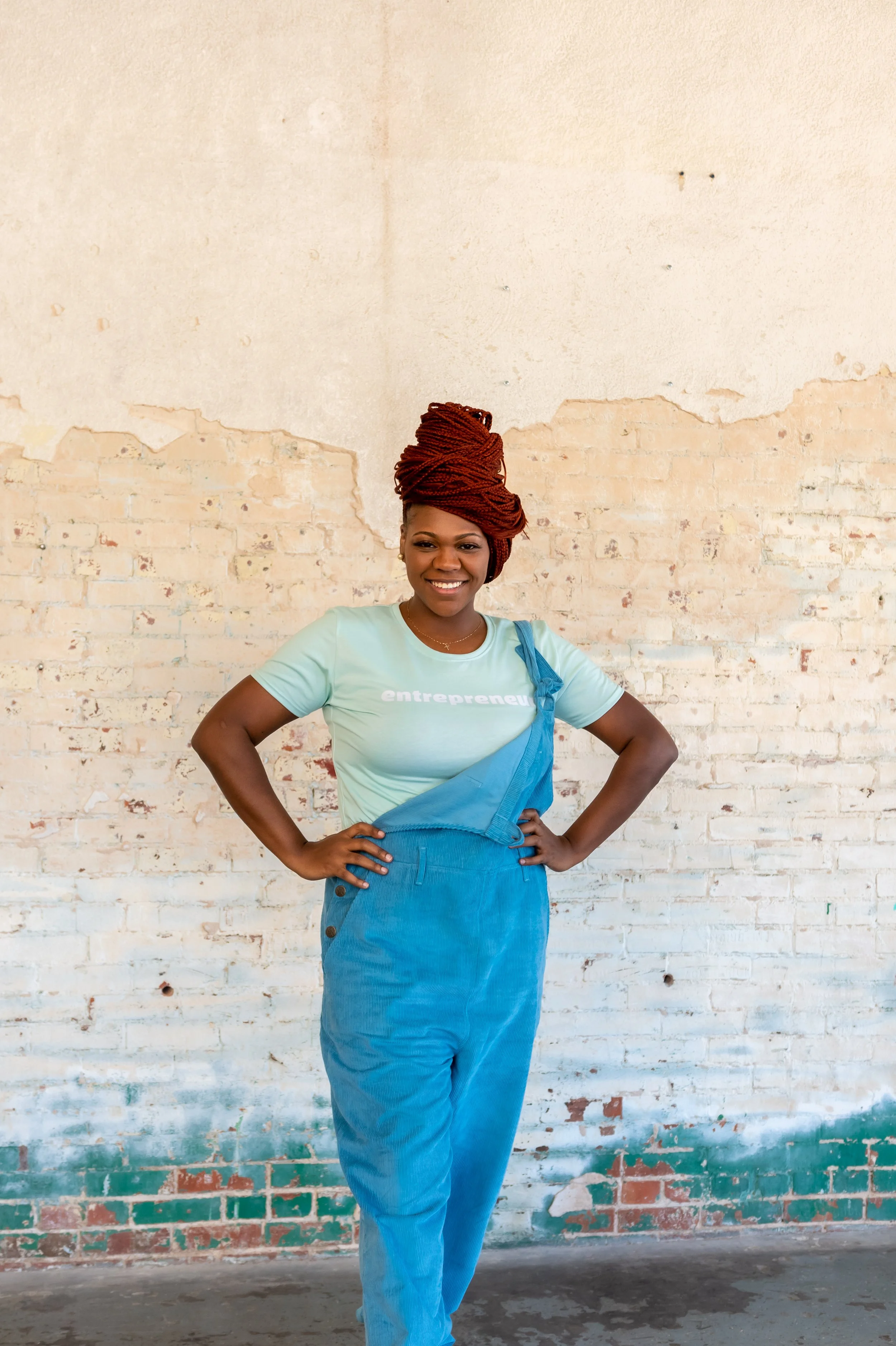 Ashley Tate with red braided updo in mint “entrepreneur” t-shirt and blue corduroy overalls, hands on hips, studio brick wall backdrop, Oklahoma City. Brand Photography.