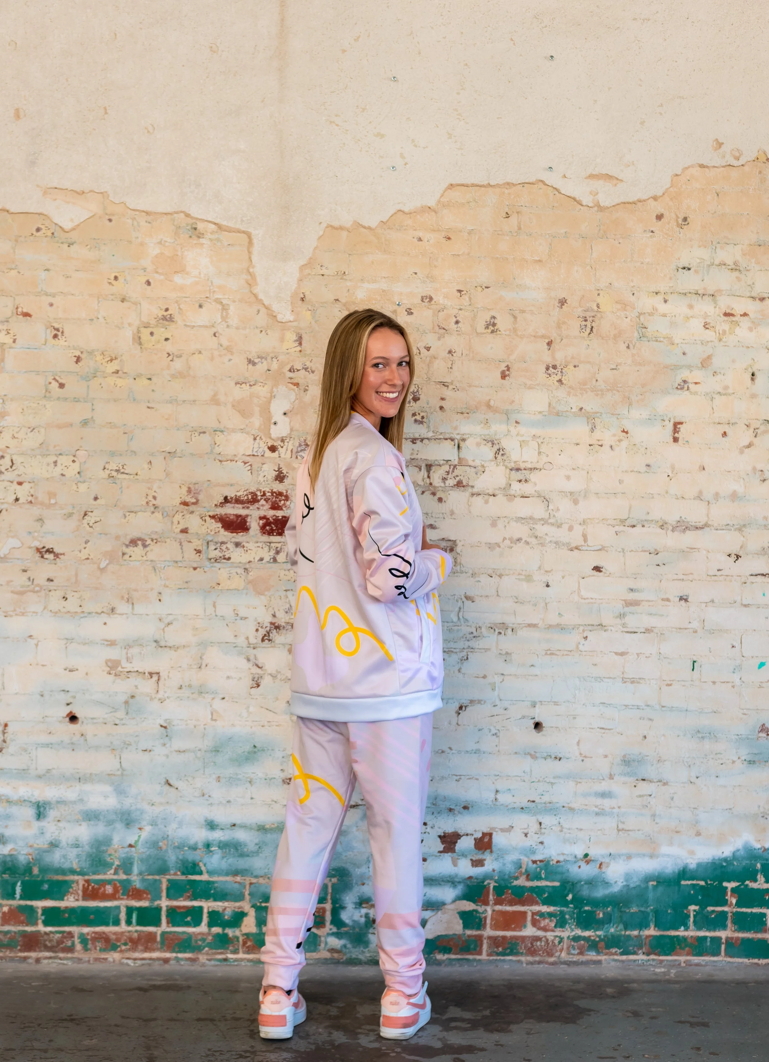 Side-profile fashion photo of blonde model wearing a pastel jacket and joggers set with yellow doodle print, sneakers, posed at a white brick wall in OKC.