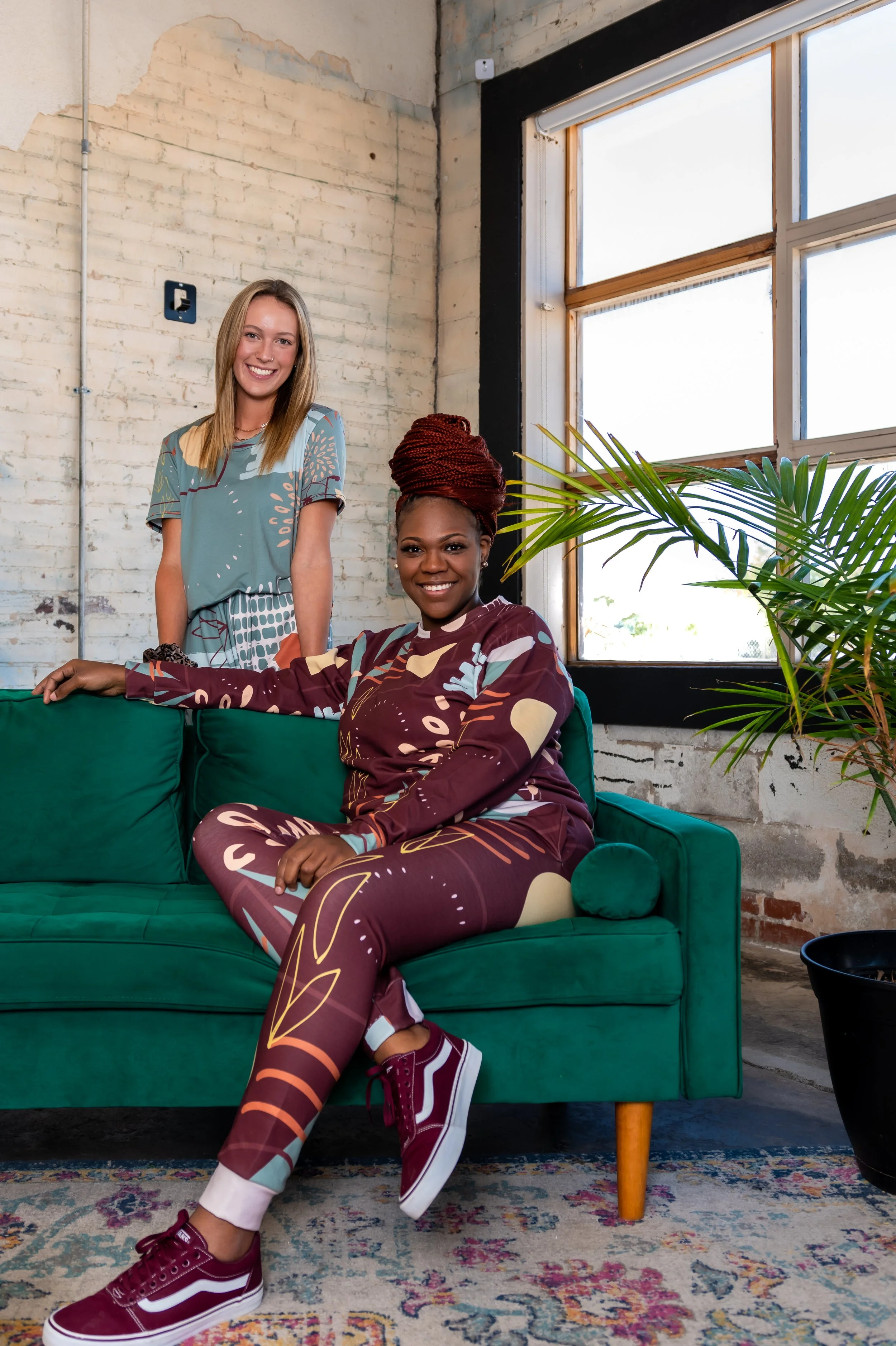 Brand portrait: Ashley Tate seated on green couch in burgundy matching set; blonde model standing behind in teal set, plants and white brick wall, Oklahoma City. Ash Bash Apparel. Brand Photography.
