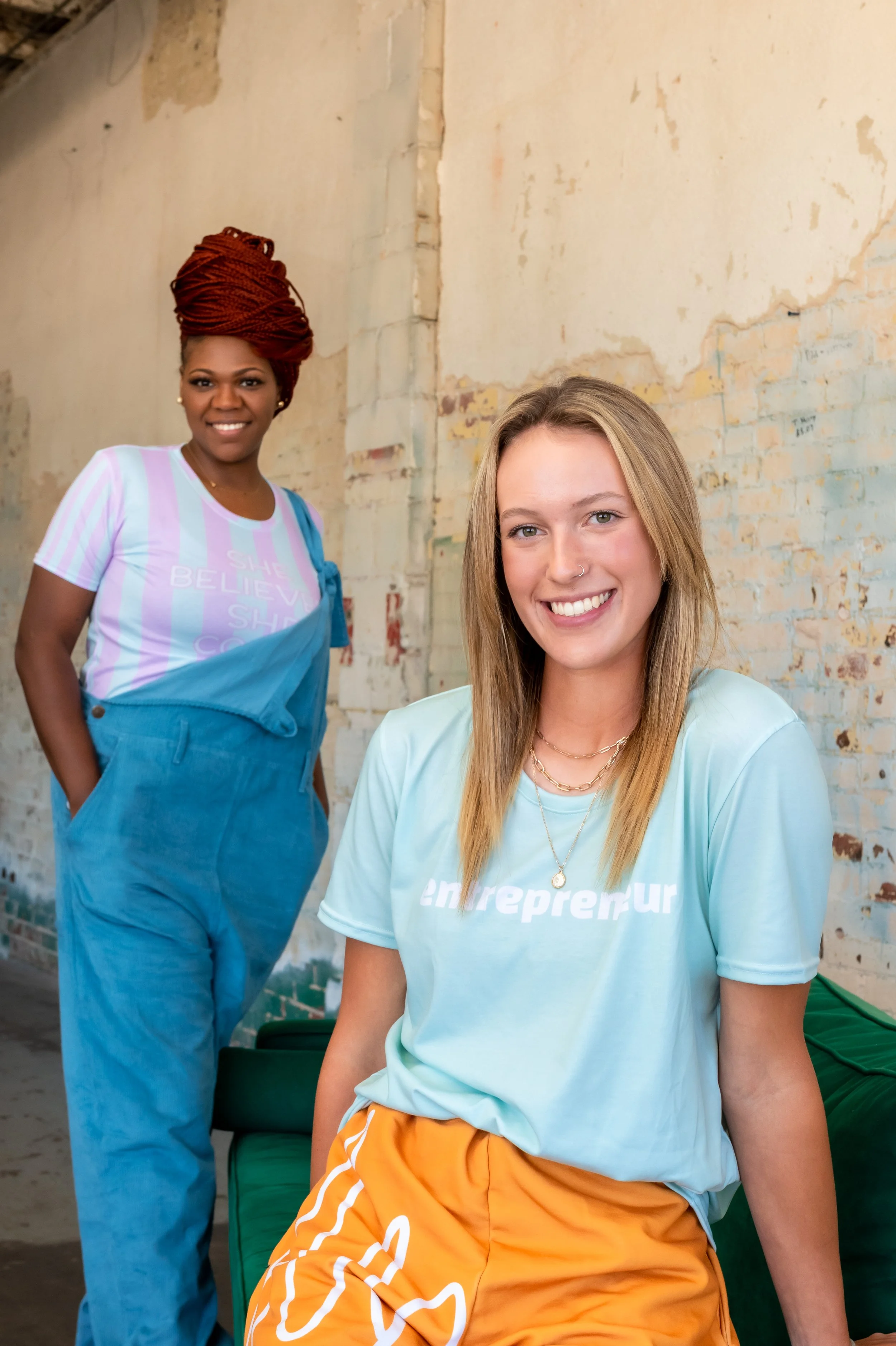 Smiling blonde model in mint “entrepreneur” t-shirt, layered gold necklaces and orange pants on green chair; Ashley Tate (red braided updo) behind in blue overalls, OKC. Brand Photography.