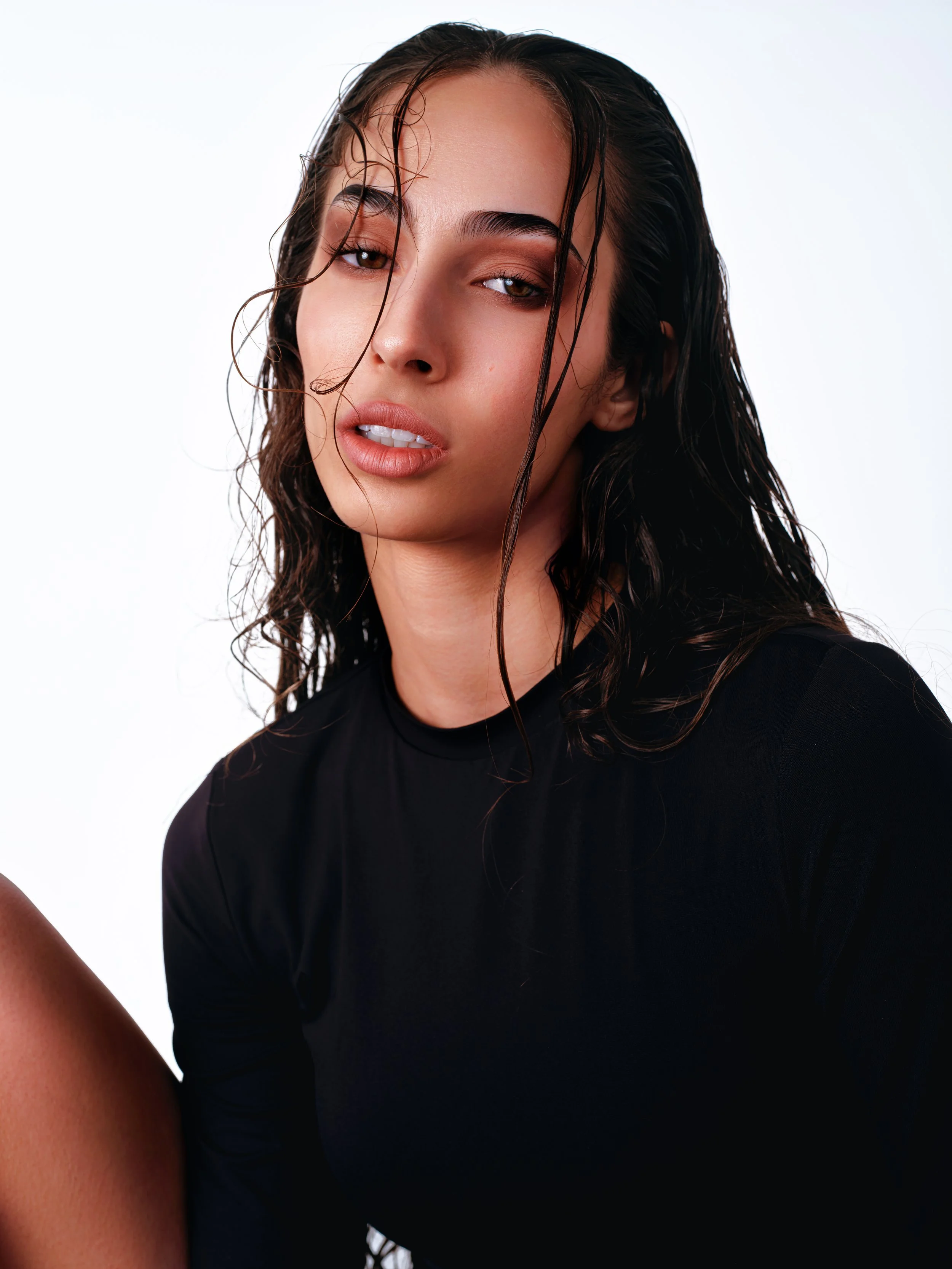 Studio editorial beauty portrait of a woman with wet-look hair and soft makeup, wearing a black top against a seamless white background.