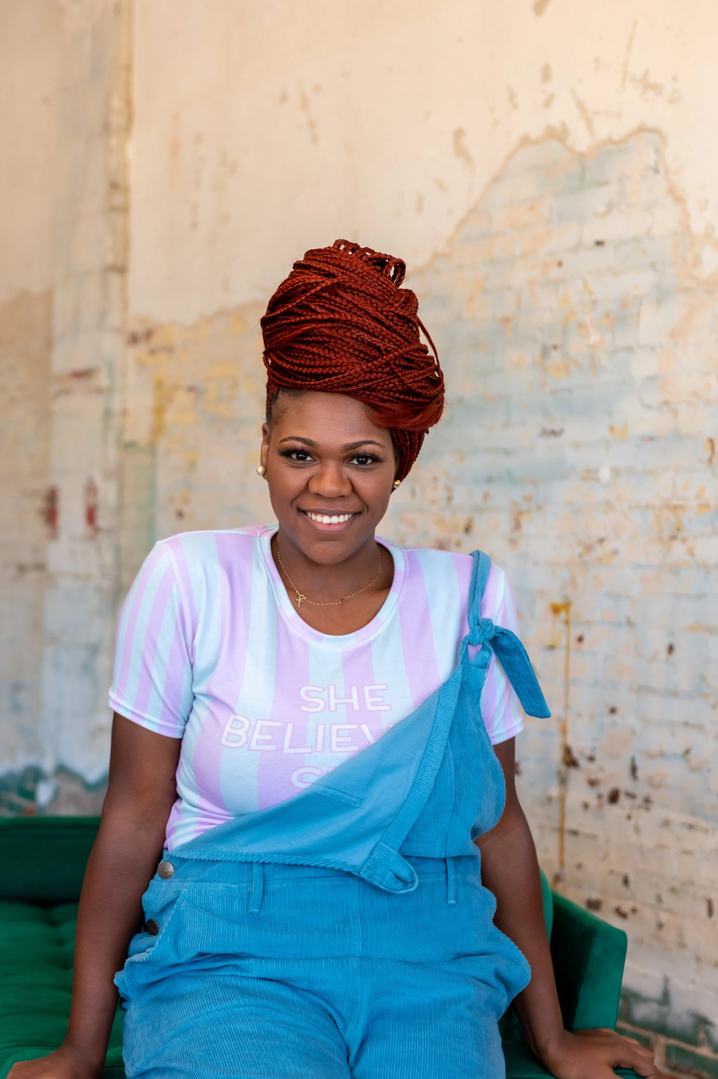 Portrait of Ashley Tate (red braided hair) in blue overalls and pastel striped “She believed…” t-shirt, gold jewelry, seated on green chair, OKC studio.