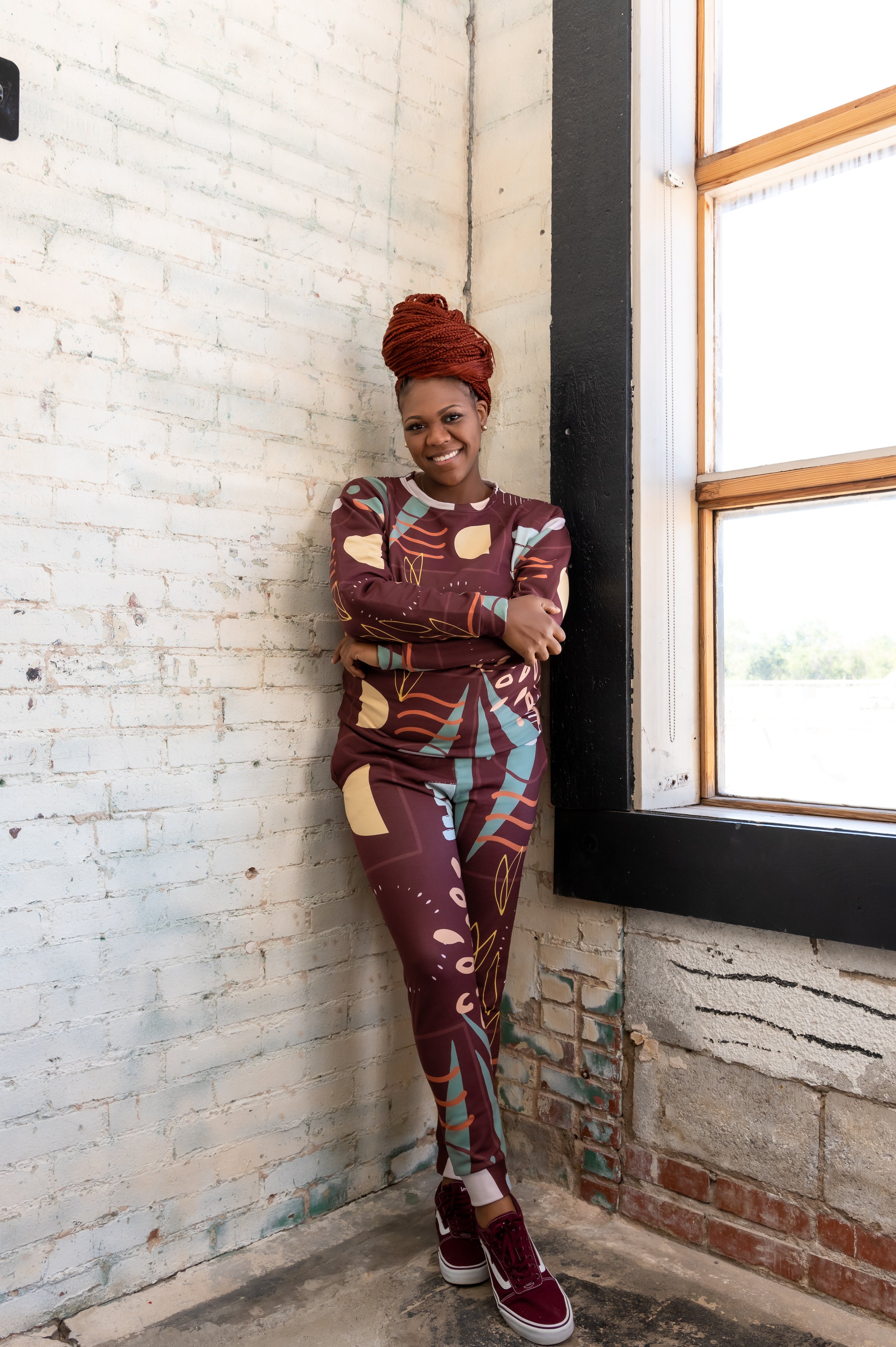Ashley Tate (red braided updo) leaning by a tall window wearing a burgundy/maroon abstract matching set and sneakers, editorial brand photo in Oklahoma City.