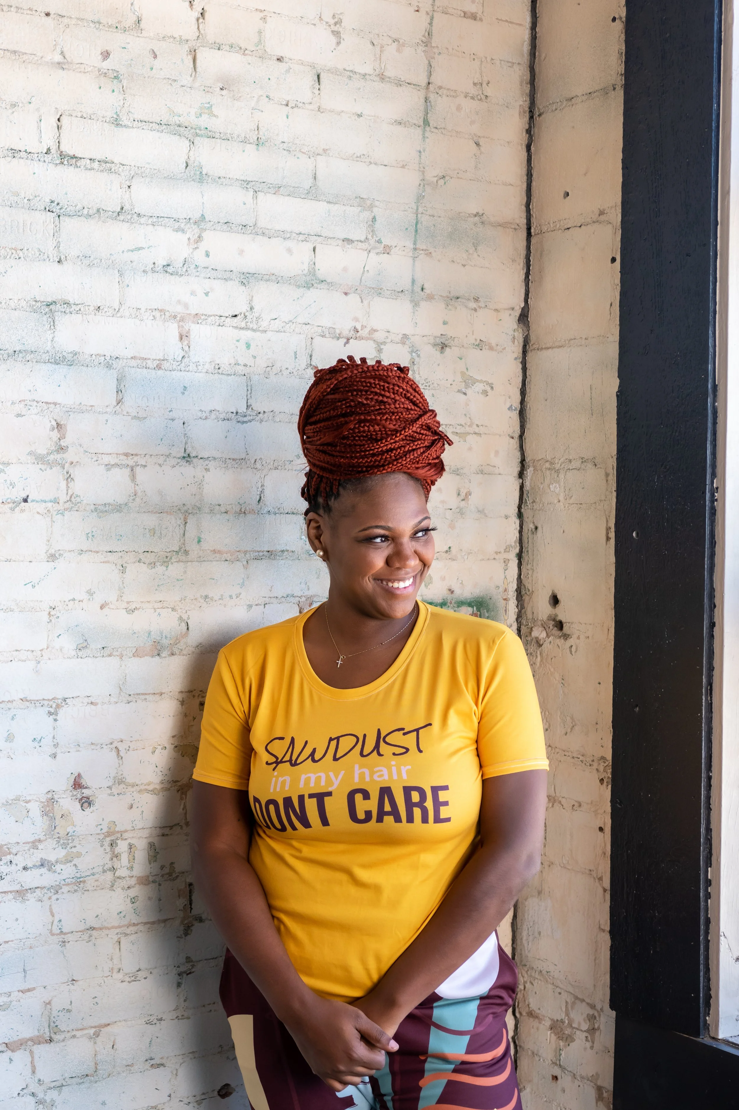 Ashley Tate (red braided hair) smiling in yellow “Sawdust in my hair don’t care” t-shirt and burgundy patterned pants, white brick wall, Oklahoma City.