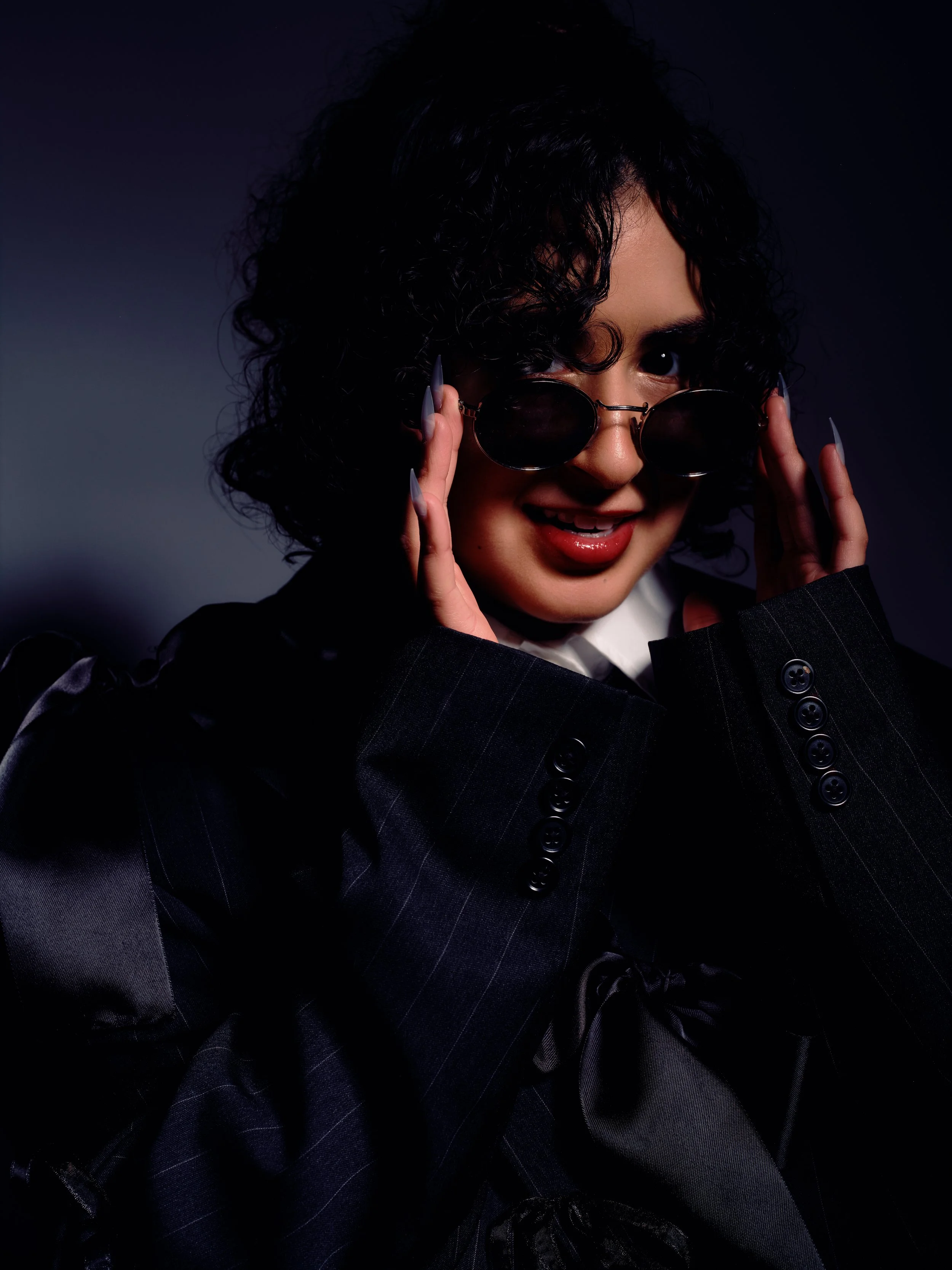 Studio editorial fashion portrait of a woman in a black pinstripe blazer and round sunglasses, smiling under dramatic shadow lighting on a dark backdrop, Oklahoma City.