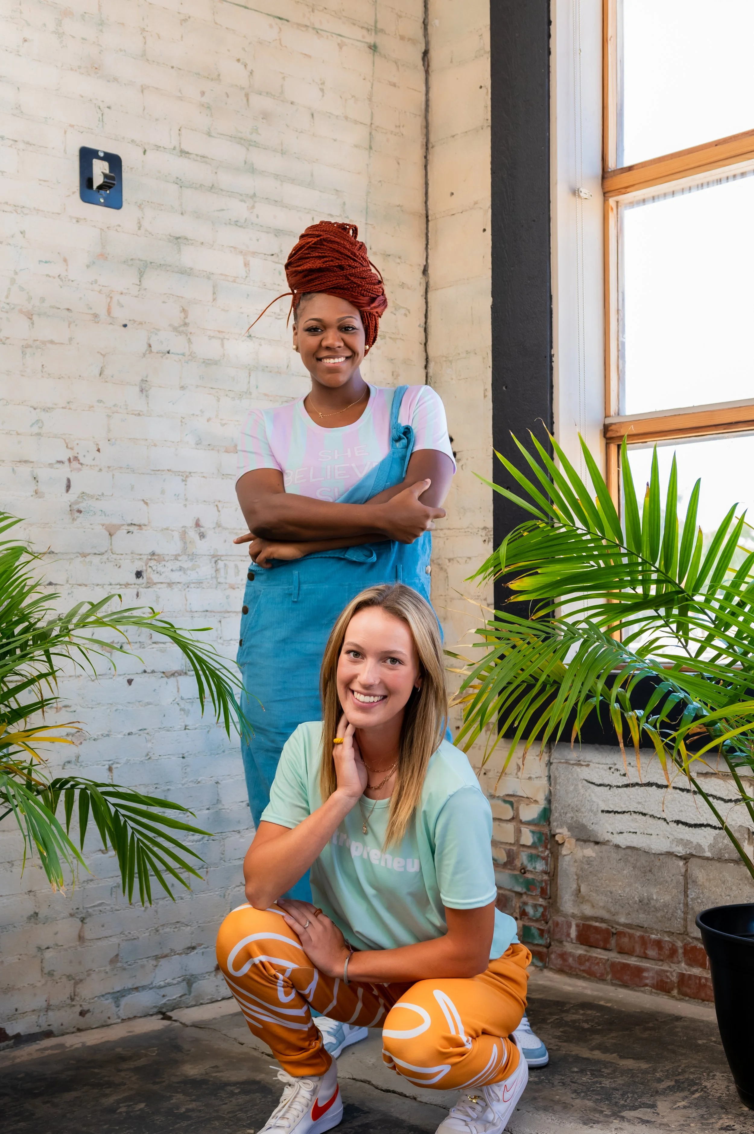 Two-model lifestyle: Ashley Tate (red braided updo) in blue overalls and striped tee standing behind a blonde model in mint tee and orange pants, plants + brick wall, OKC. Brand Photography.