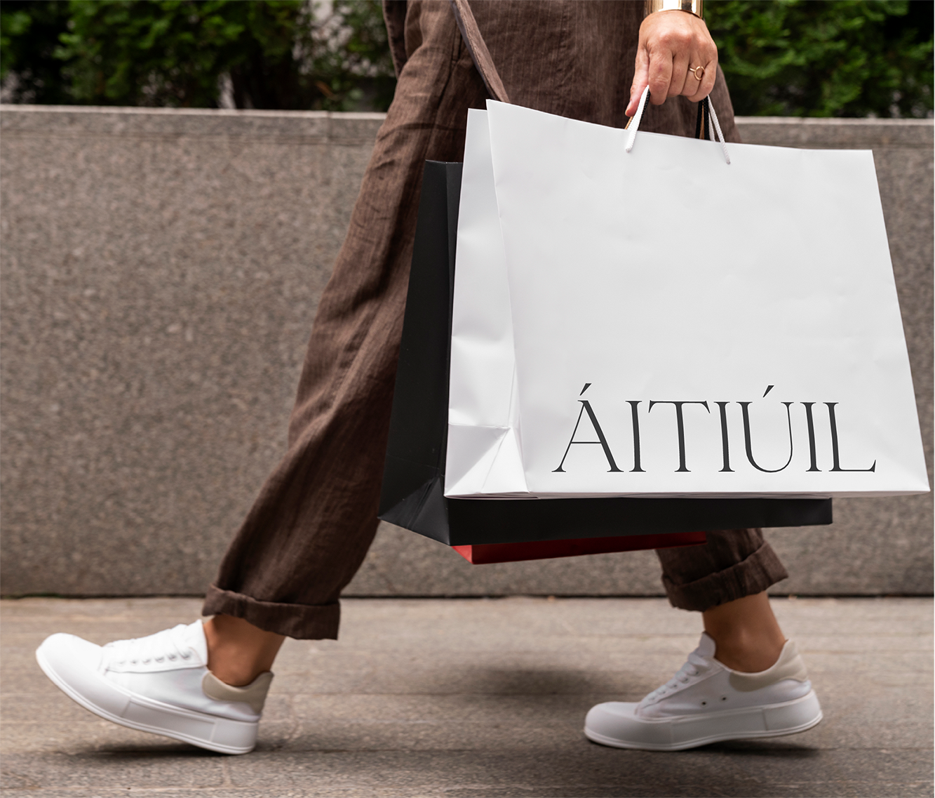 Person walking outdoors carrying a large white ÁITIÚIL shopping bag with serif logo, alongside a black bag with a red interior lining, wearing brown wide-leg trousers and white sneakers.