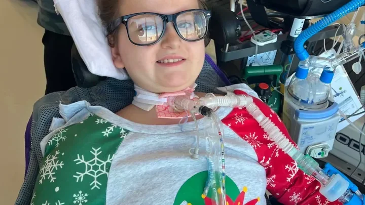 A young girl in a hospital bed wearing glasses and a holiday-themed shirt with snowflakes, connected to medical machines and tubes.