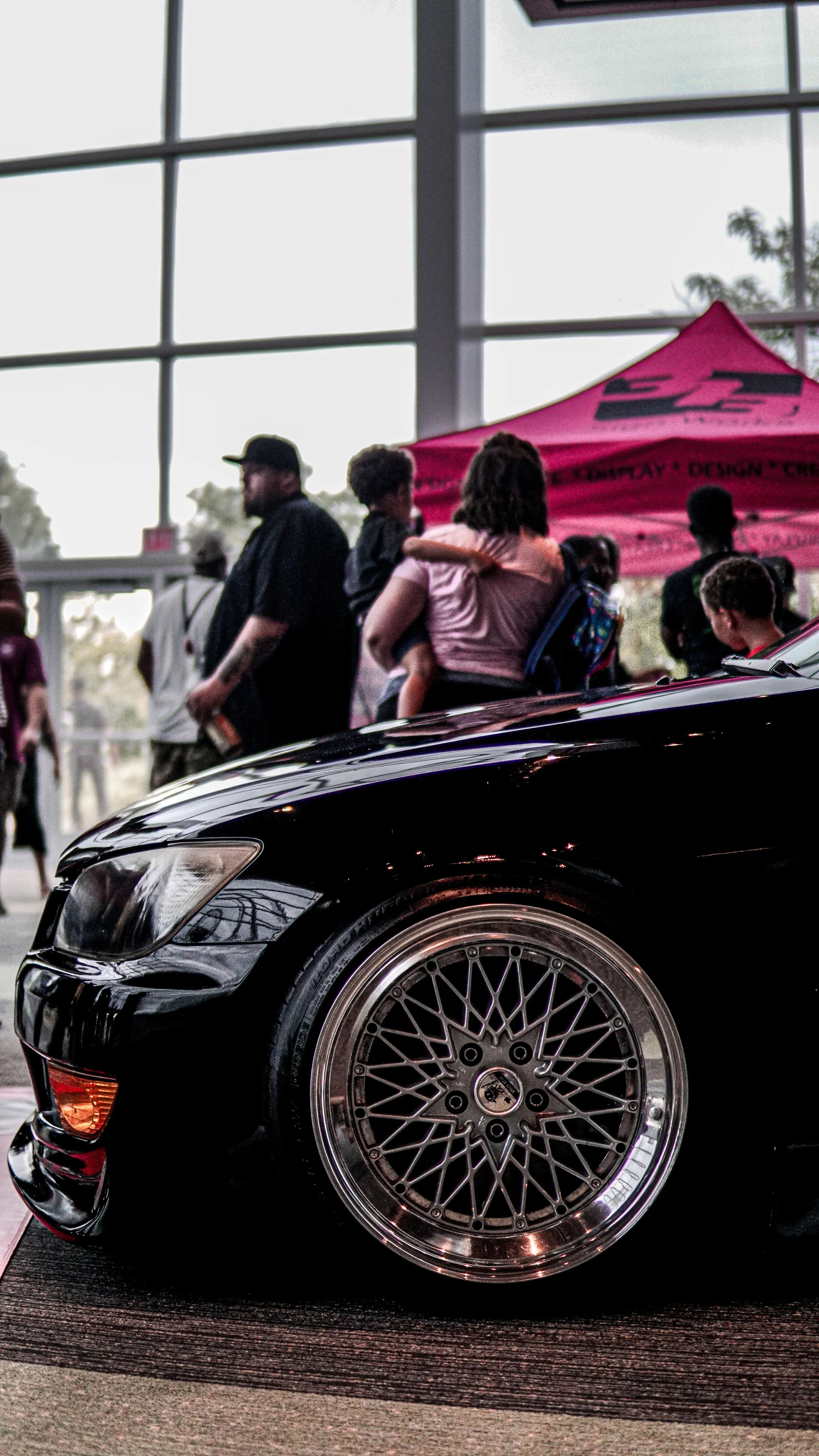 Close-up of the front of a black car with custom alloy wheels displayed at an indoor auto show, with people and a pink tent in the background.