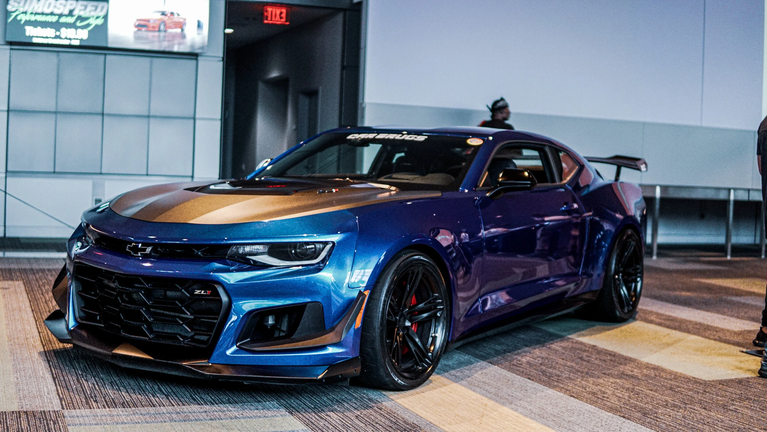 A blue Chevrolet Camaro ZL1 sports car displayed indoors, with a gold stripe on its hood, black wheels, and a rear wing.