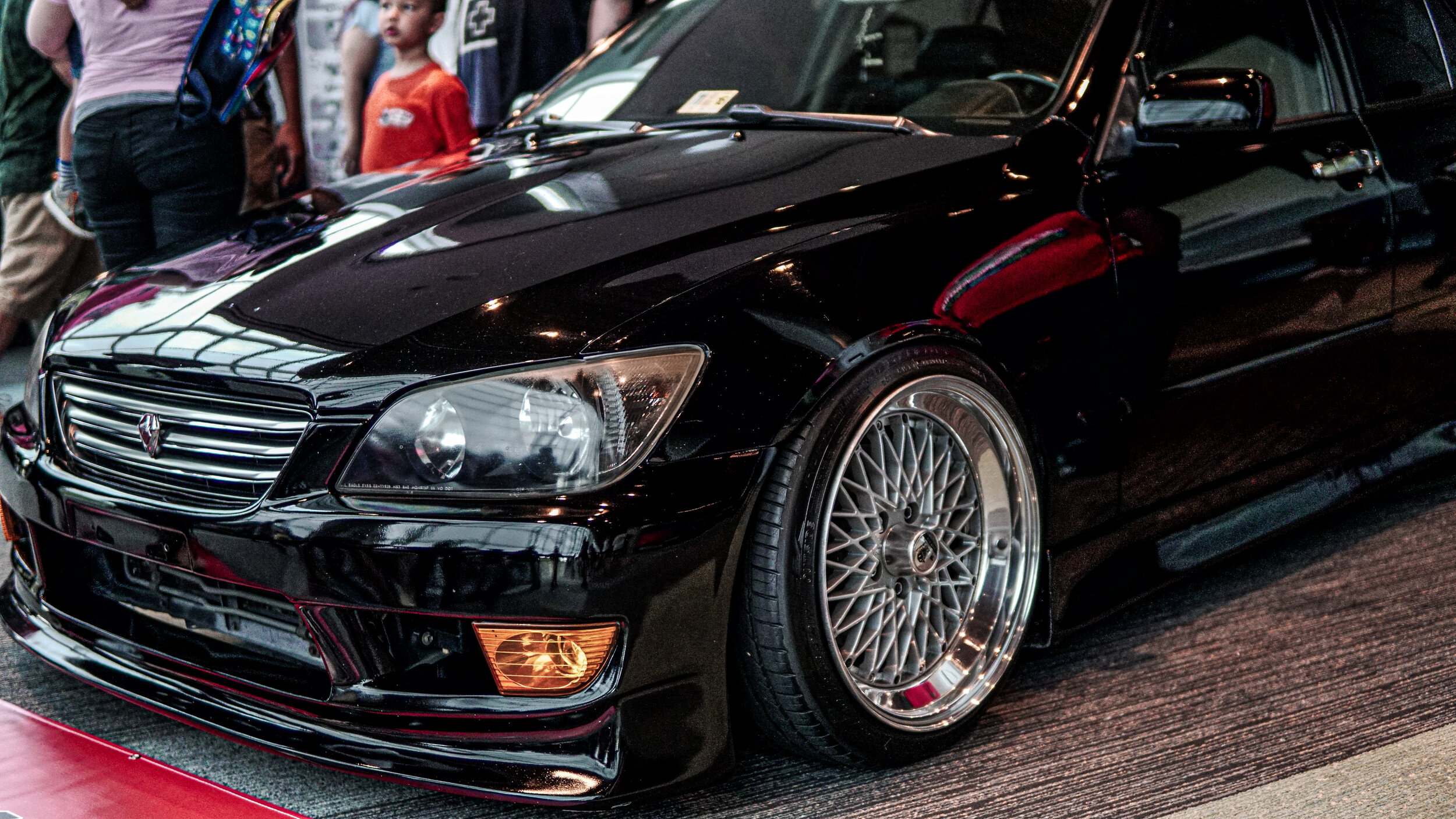 Black luxury sedan on display at an auto show, with people in the background