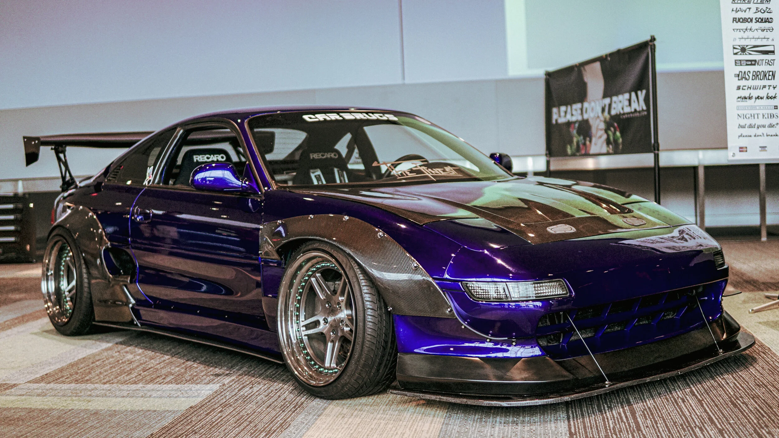 A purple custom sports car with carbon fiber fenders, wide tires, and a large rear wing on display indoors, with banners in the background.