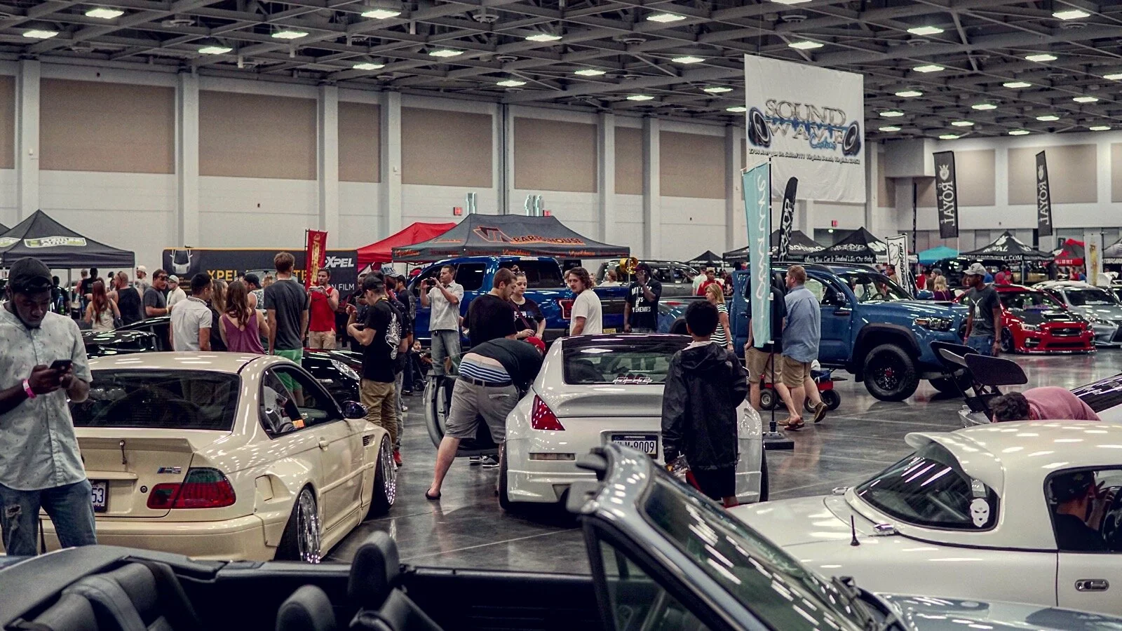 Indoor car show with various cars on display and many attendees looking at vehicles, tents, and banners.