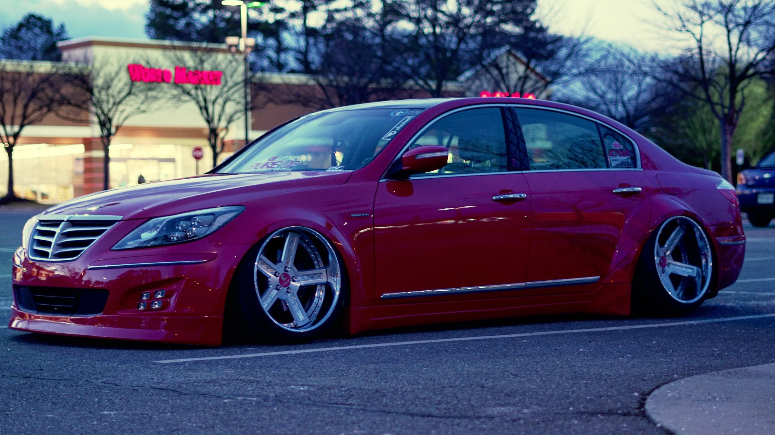 A lowered, customized red sedan with large chrome wheels parked in a parking lot in front of a store with a sign that reads 'WONCO MART.' Trees without leaves are visible in the background, indicating it is likely late fall or winter.