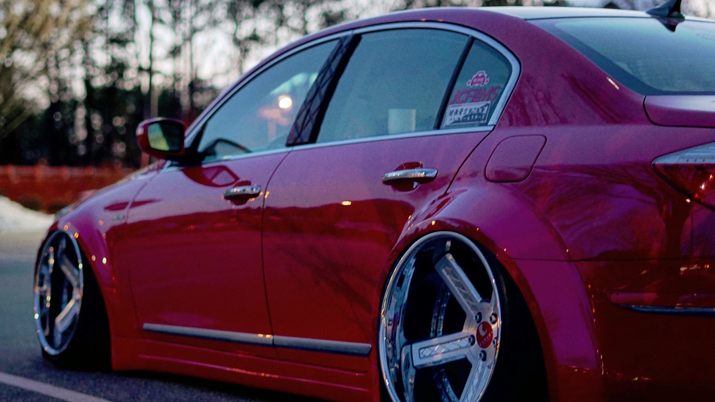 Red sports car parked outdoors during dusk with large custom rims and a sticker on the rear side window.