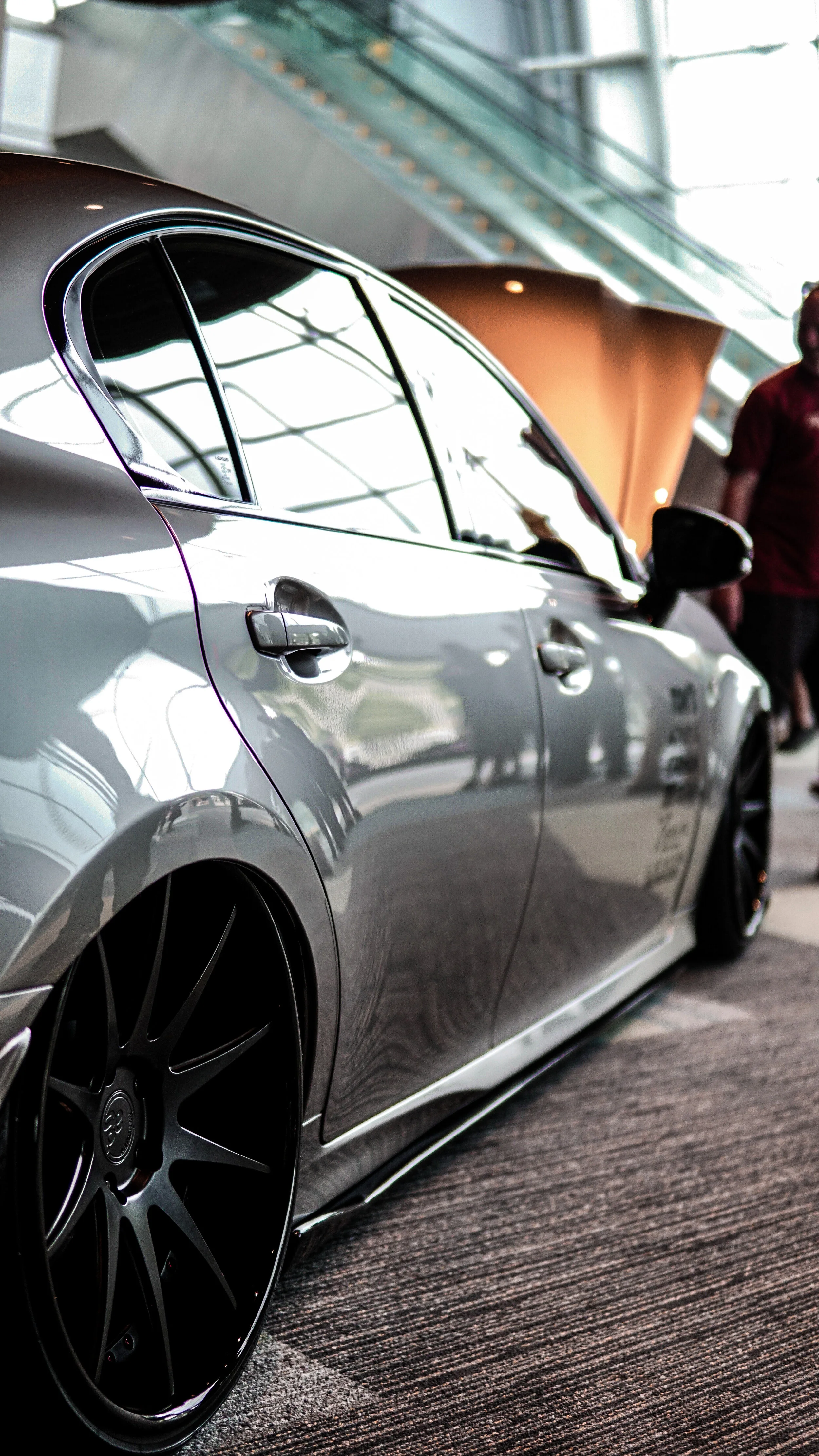 Close-up of a silver luxury car with tinted windows and black wheels, displayed indoors at a car show.