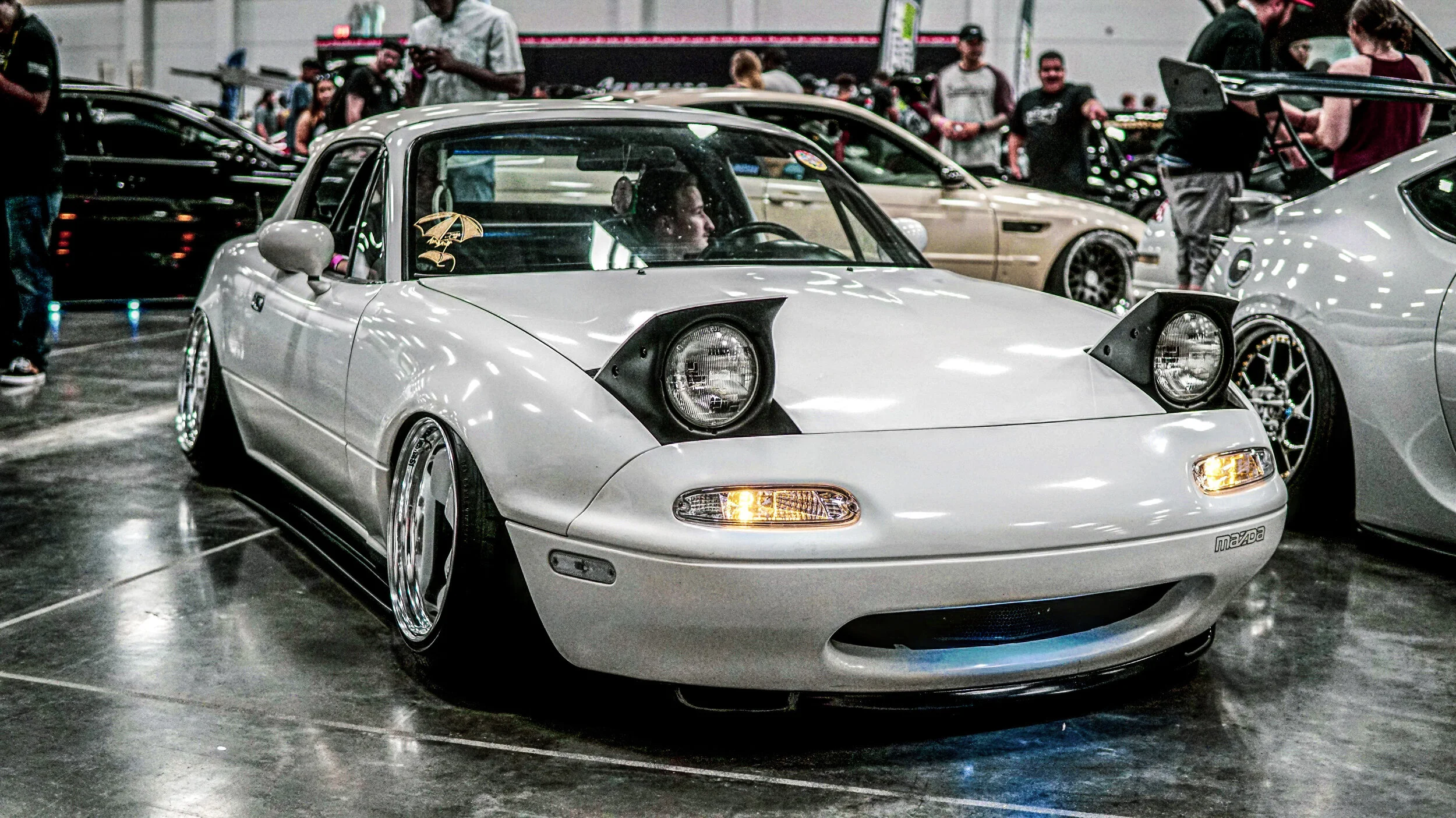 A customized Mazda MX-5 Miata with pop-up headlights at a car show, surrounded by other cars and attendees.