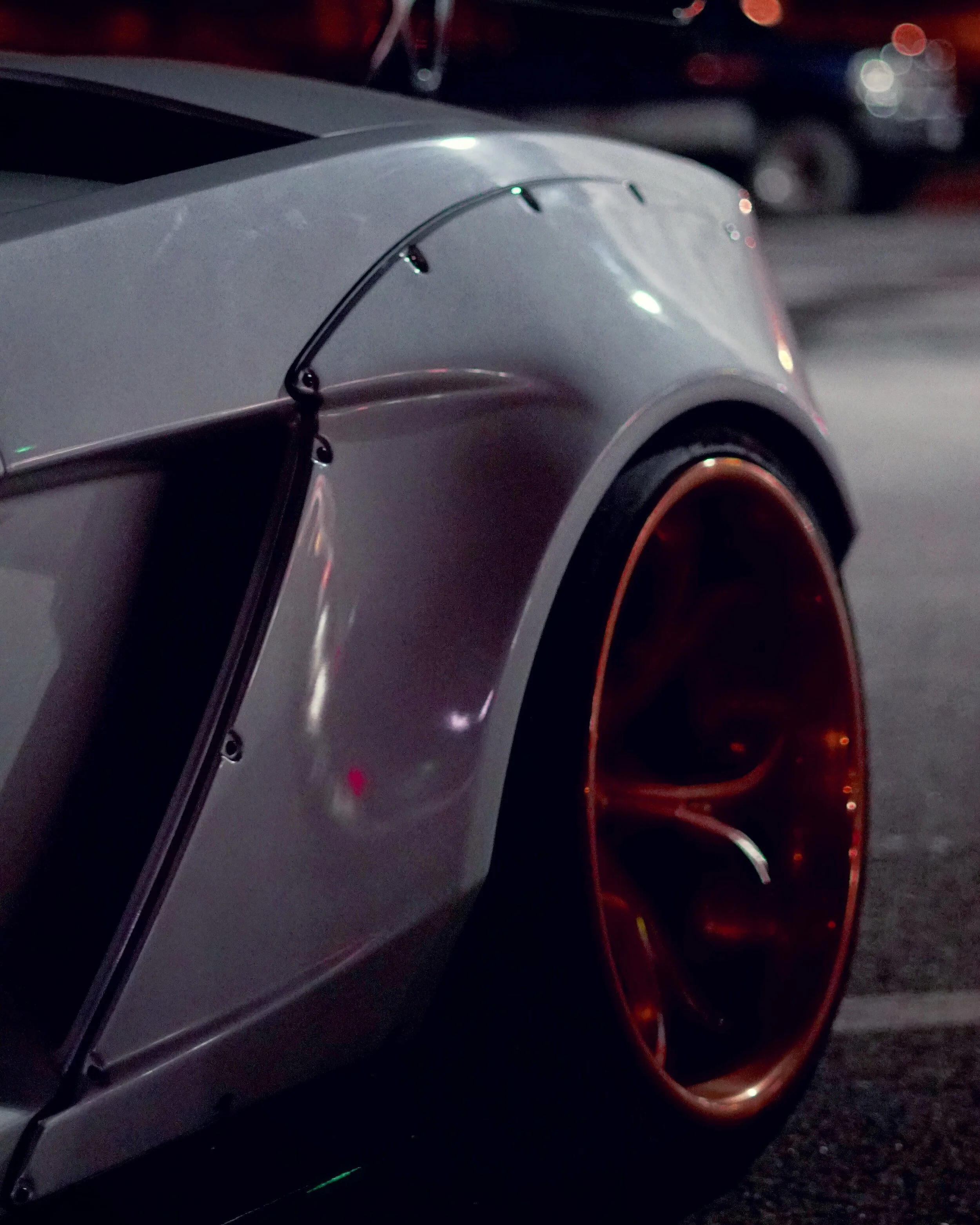 Close-up of a metallic silver sports car with a red-orange rim on the tire, parked at night.