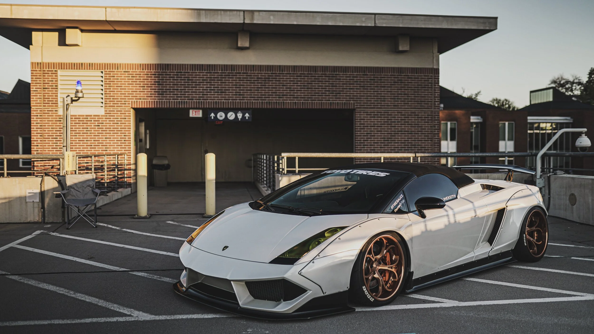 White and black Lamborghini sports car with bronze rims parked in a parking lot near a brick building