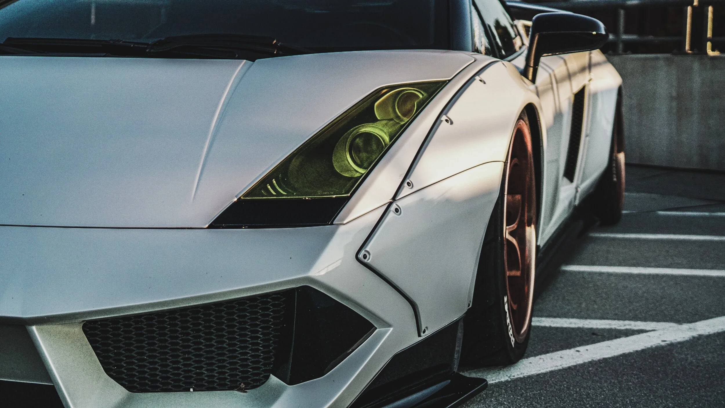 A white sports car with custom modifications, including a wide body kit, bronze wheels, and green-tinted headlights, parked in a parking lot.